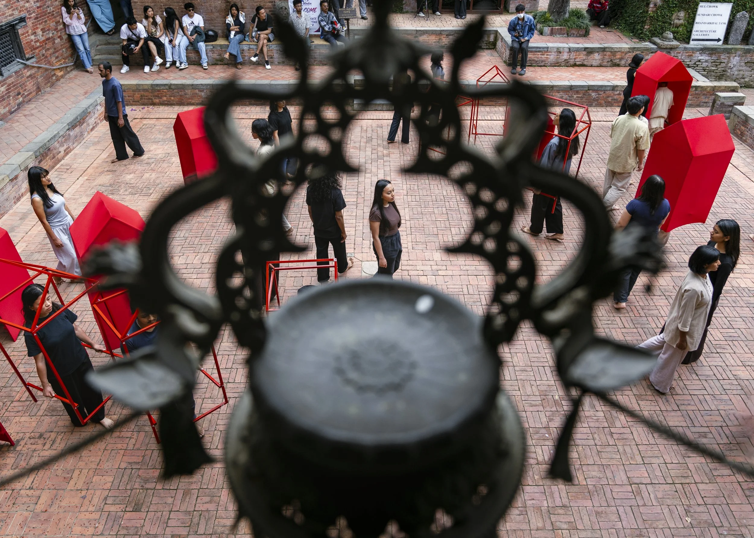 People sitting on benches and walking in an outdoor brick courtyard, viewed through a decorative black metal fence with a blurred foreground.