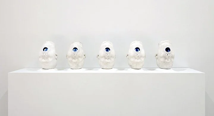 Five white sculpture heads with large blue eyes on a white pedestal against a plain white wall.
