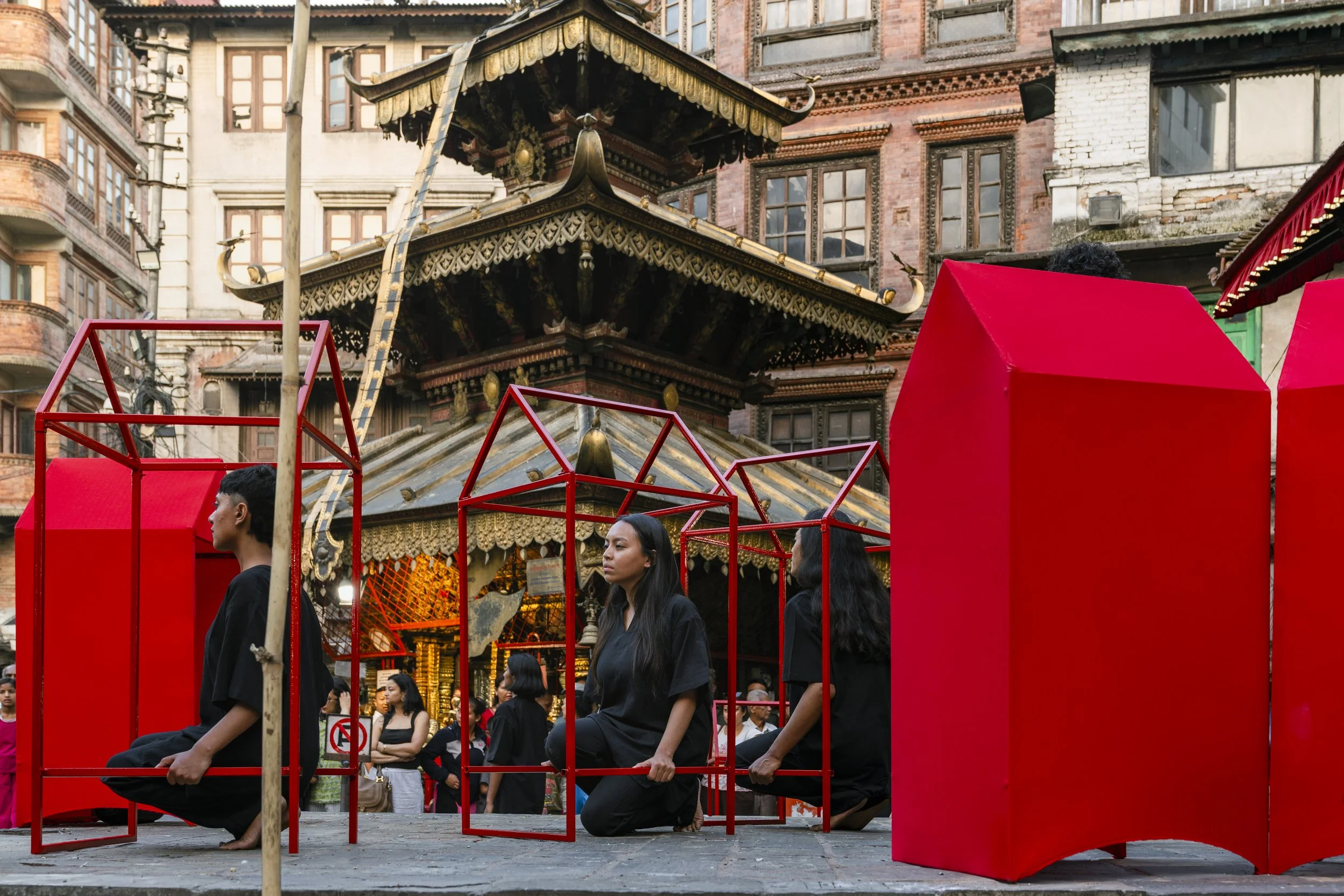 People sitting on red chairs in front of a traditional Asian temple structure, with many onlookers in the background.