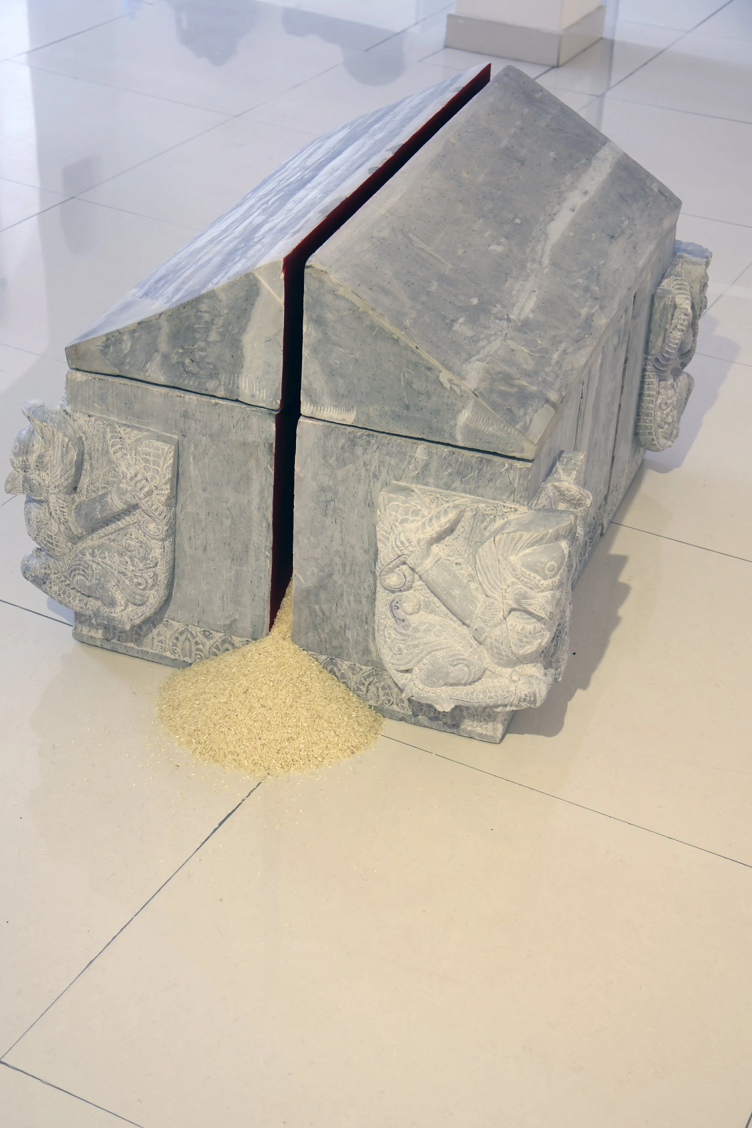 A stone sculpture resembling a house with carved decorative elements on the sides, topped with a geometric roof, with a small pile of rice or grains spilled underneath.