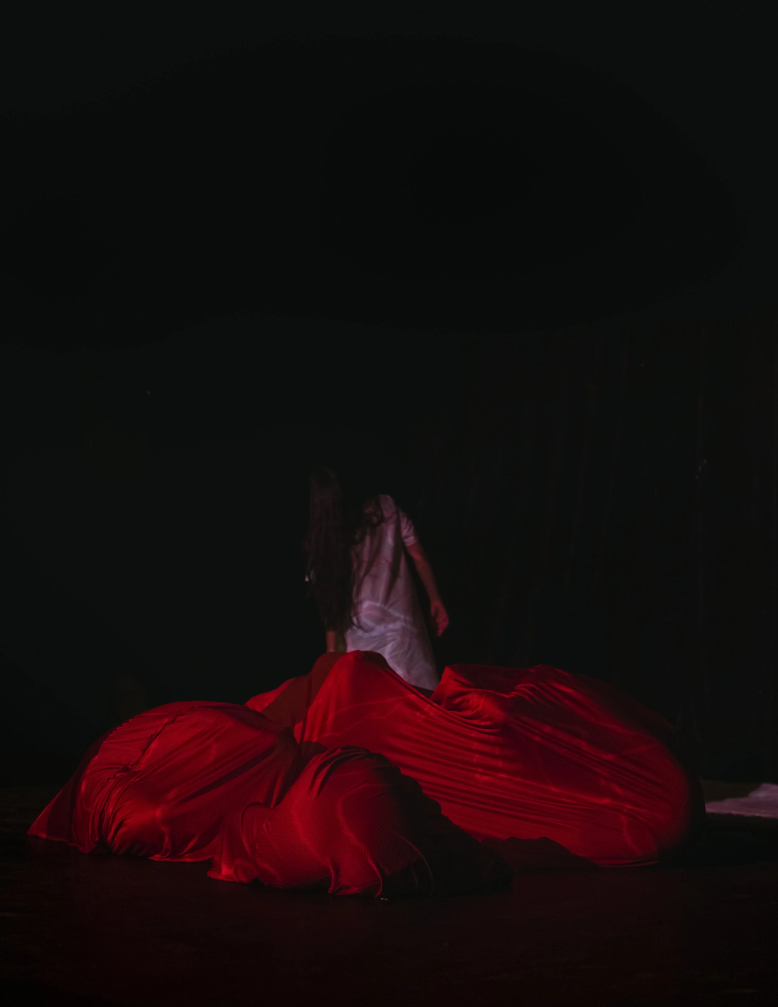 Person wearing a pink jacket and engaged in a dance or performance with red fabric, illuminated by red lighting against a dark background.