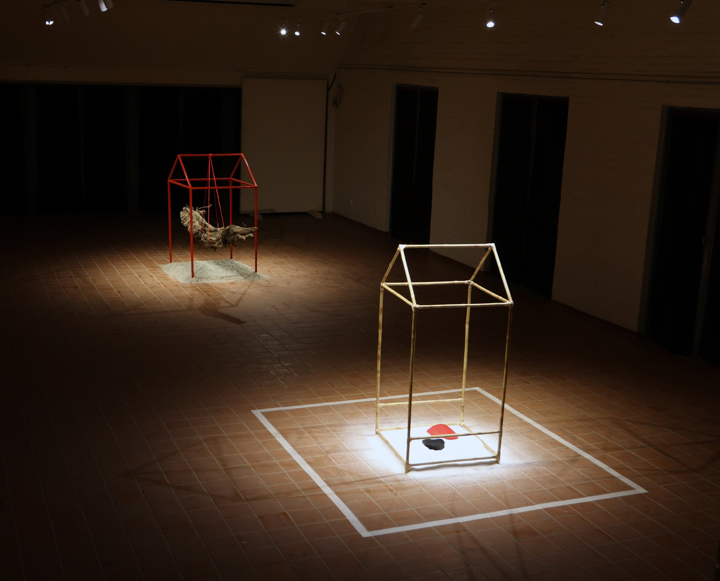 An art installation featuring two geometric sculptures on a tiled floor in a dimly lit gallery space. One sculpture has a red frame with a swinging figure inside, while the other is an empty beige frame.