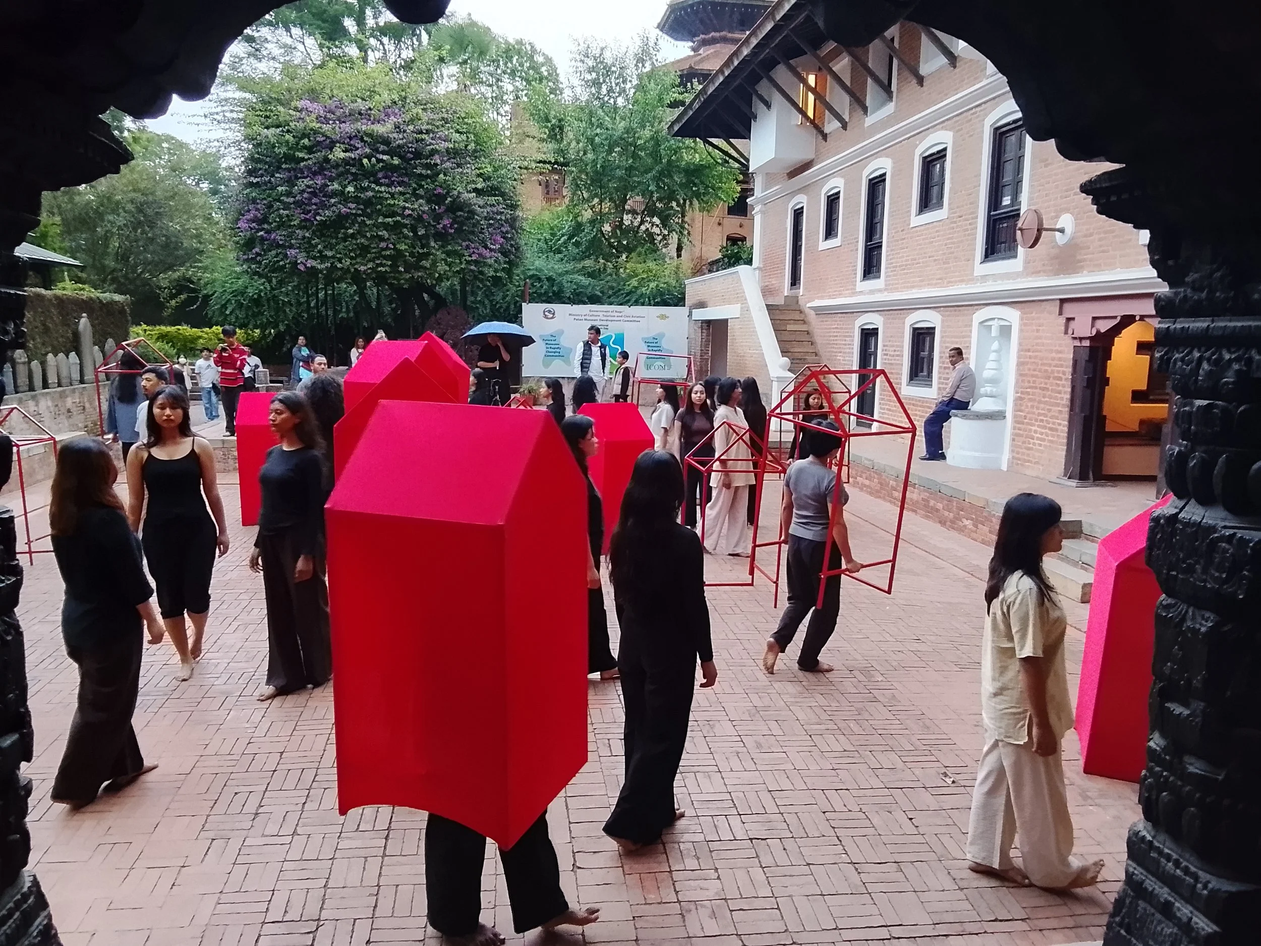 People walking and standing in an outdoor courtyard area with trees, a brick building, and modern art sculptures in bright pink. There is a mix of men and women, some holding umbrellas, and a few looking at the camera.
