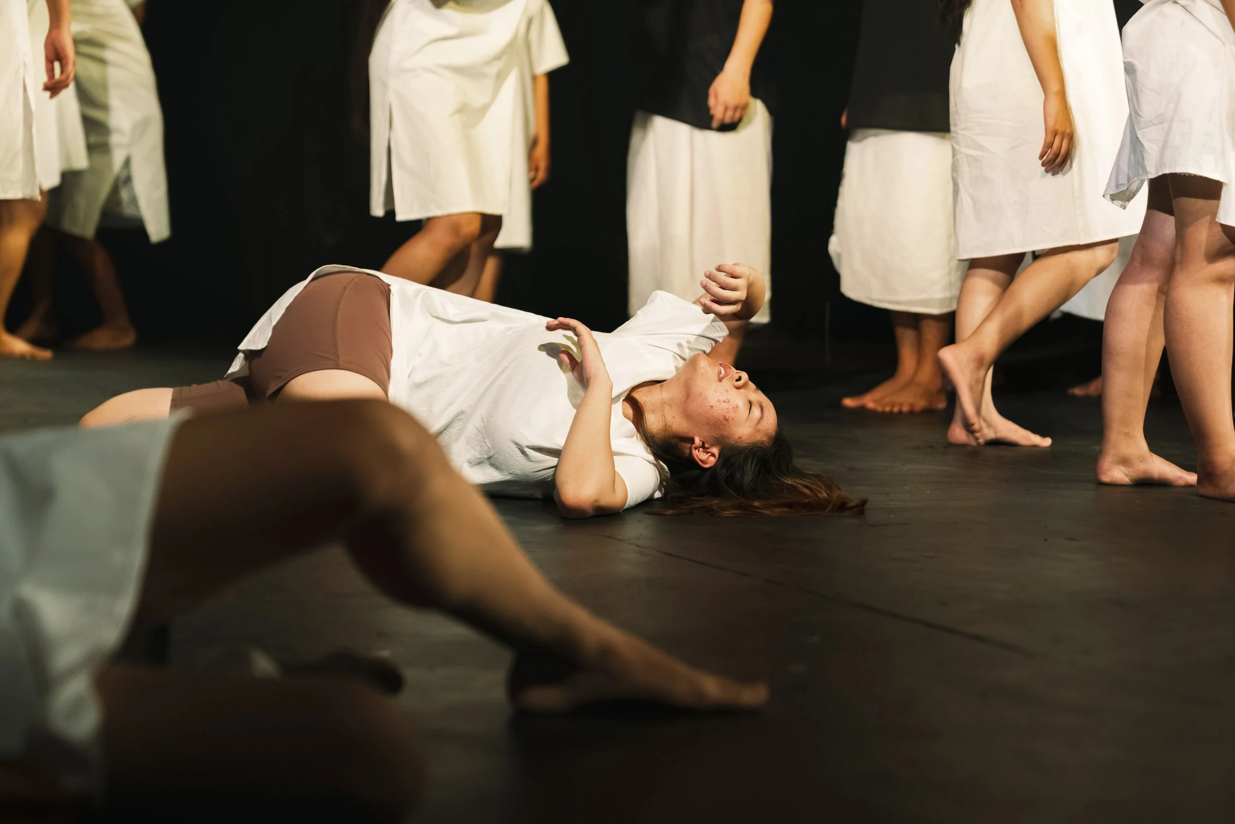 A person lying on the floor with eyes closed, surrounded by people standing around in white robes, possibly during a theatrical or artistic performance.