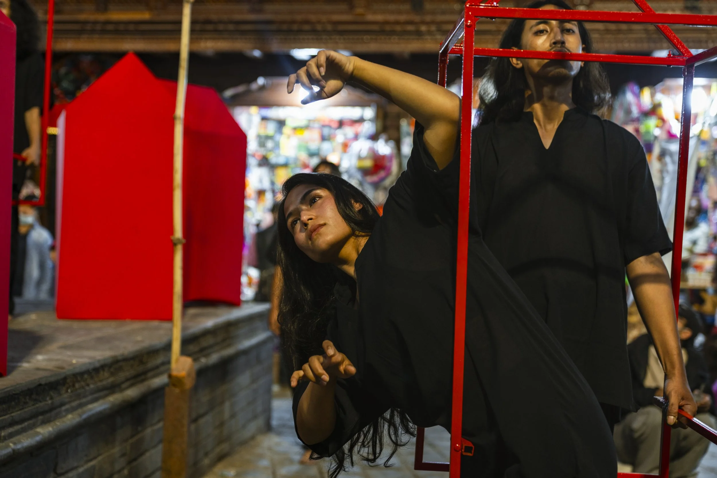 Two women with long dark hair, dressed in black, are performing a dance or pose behind a red geometric frame in a colorful indoor market or event space. One woman is leaning back and the other is standing upright, with the background filled with blur