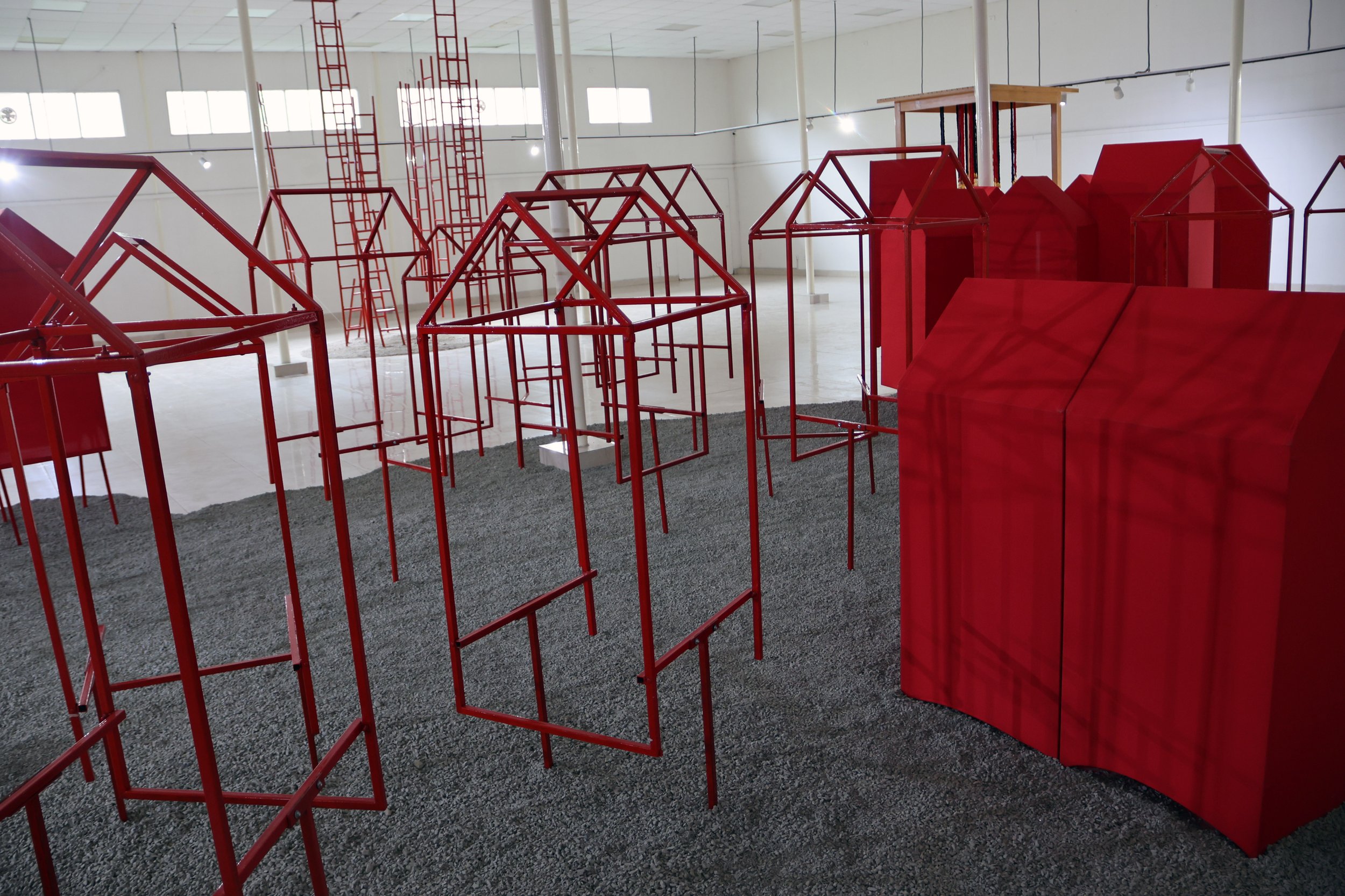 Indoor art installation featuring red metal frames shaped like small houses and structures on a gray carpeted floor.