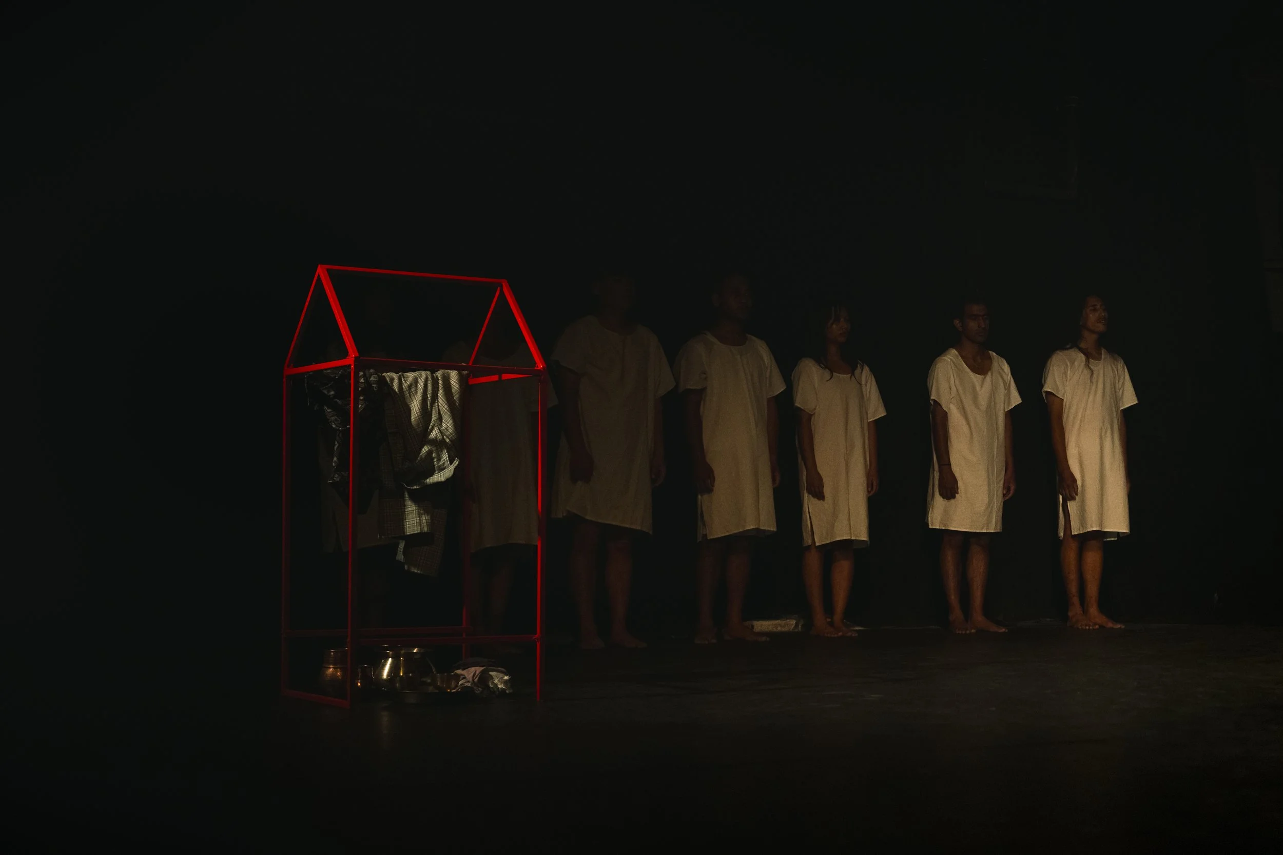 People dressed in white standing in a line in a dark stage, with an illuminated red geometric structure on the left side of the image.