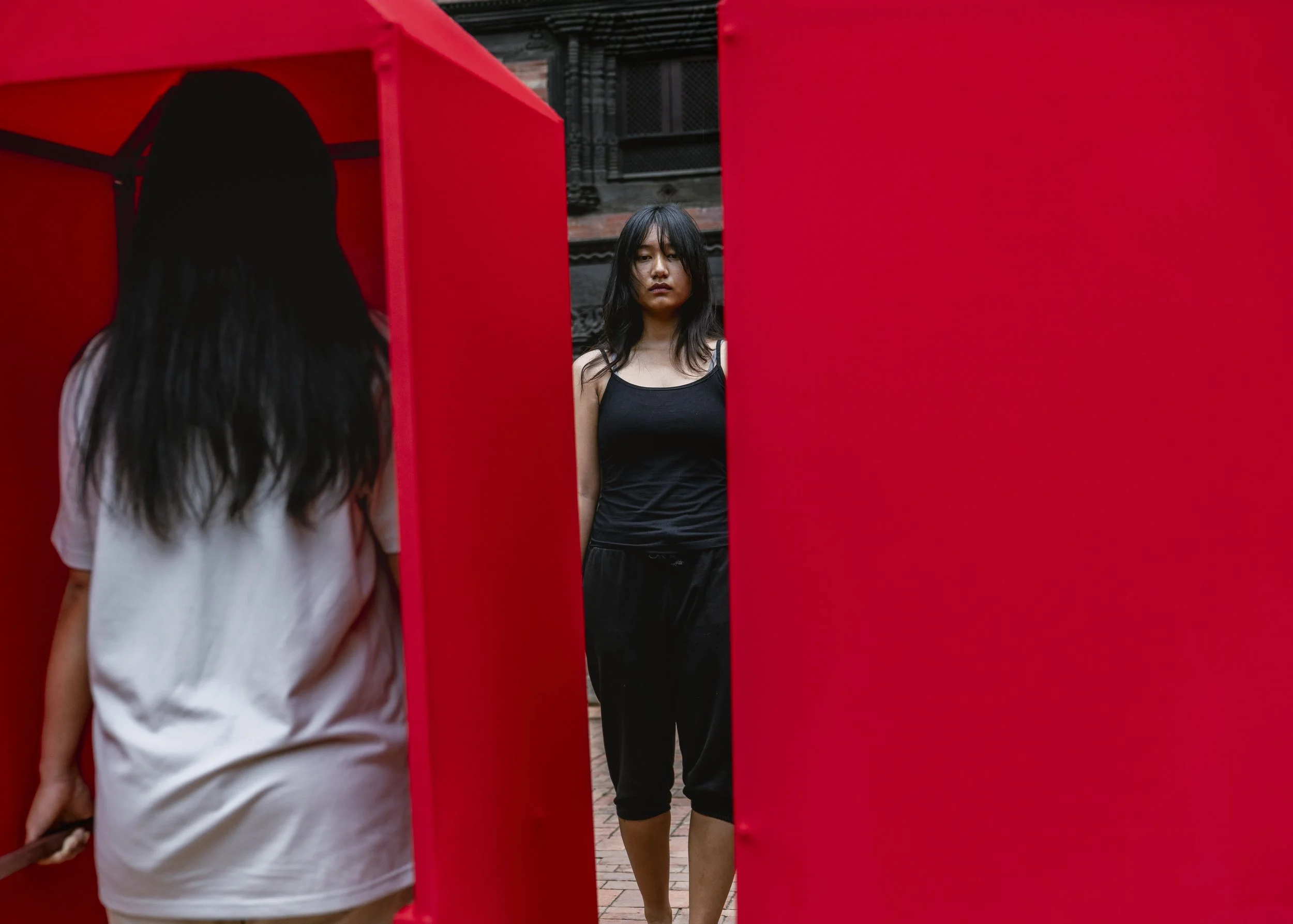 A woman with long black hair wearing a black tank top and black pants looking at her reflection in a mirror. She is surrounded by red panels, with a person in a white shirt visible in the foreground. The background features a brick building and metal