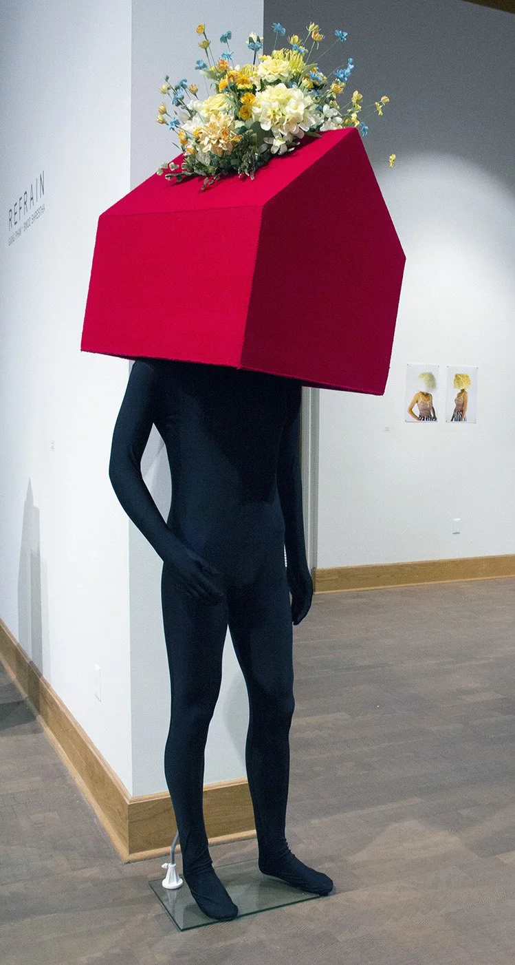 A mannequin with a black bodysuit and black shoes, with a large red cube-shaped container on its head, filled with colorful flowers.
