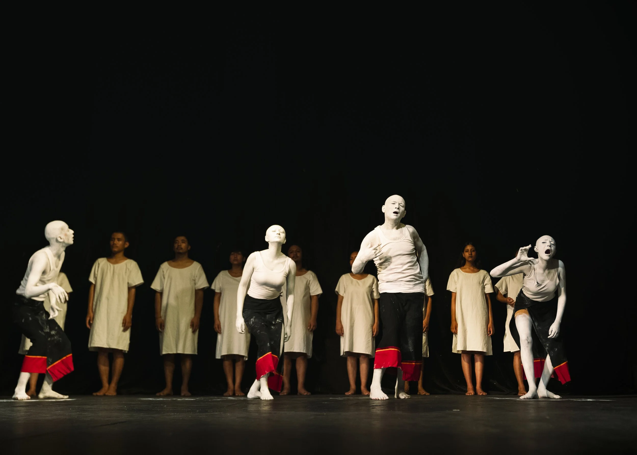 Contemporary theater performers in white makeup and black-and-red costumes with a chorus on a dark stage