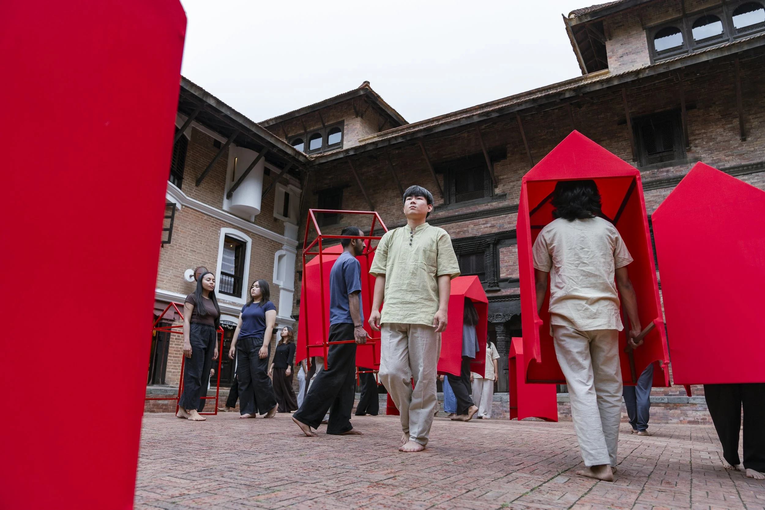 People participating in an outdoor performance or installation, with some standing in red geometric structures, in a courtyard with brick buildings in the background.