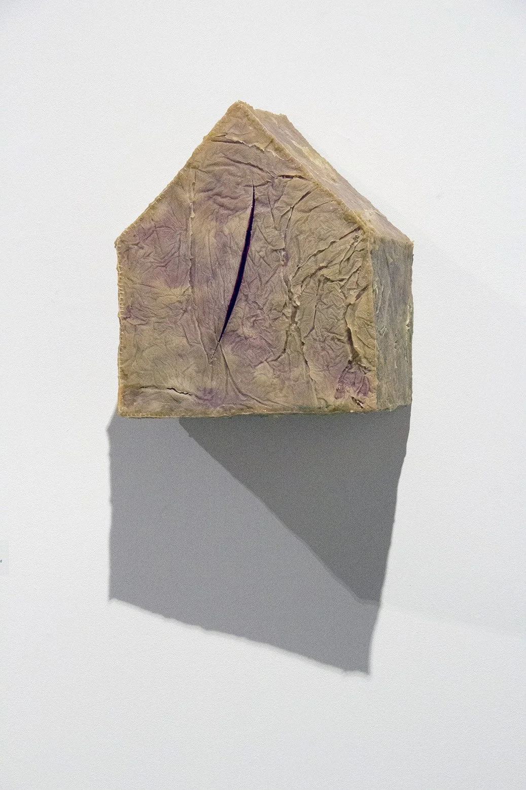 A rectangular stone with a pointed top, resembling a miniature house or monument, with a crack running through its center, cast in shadow on a white background.