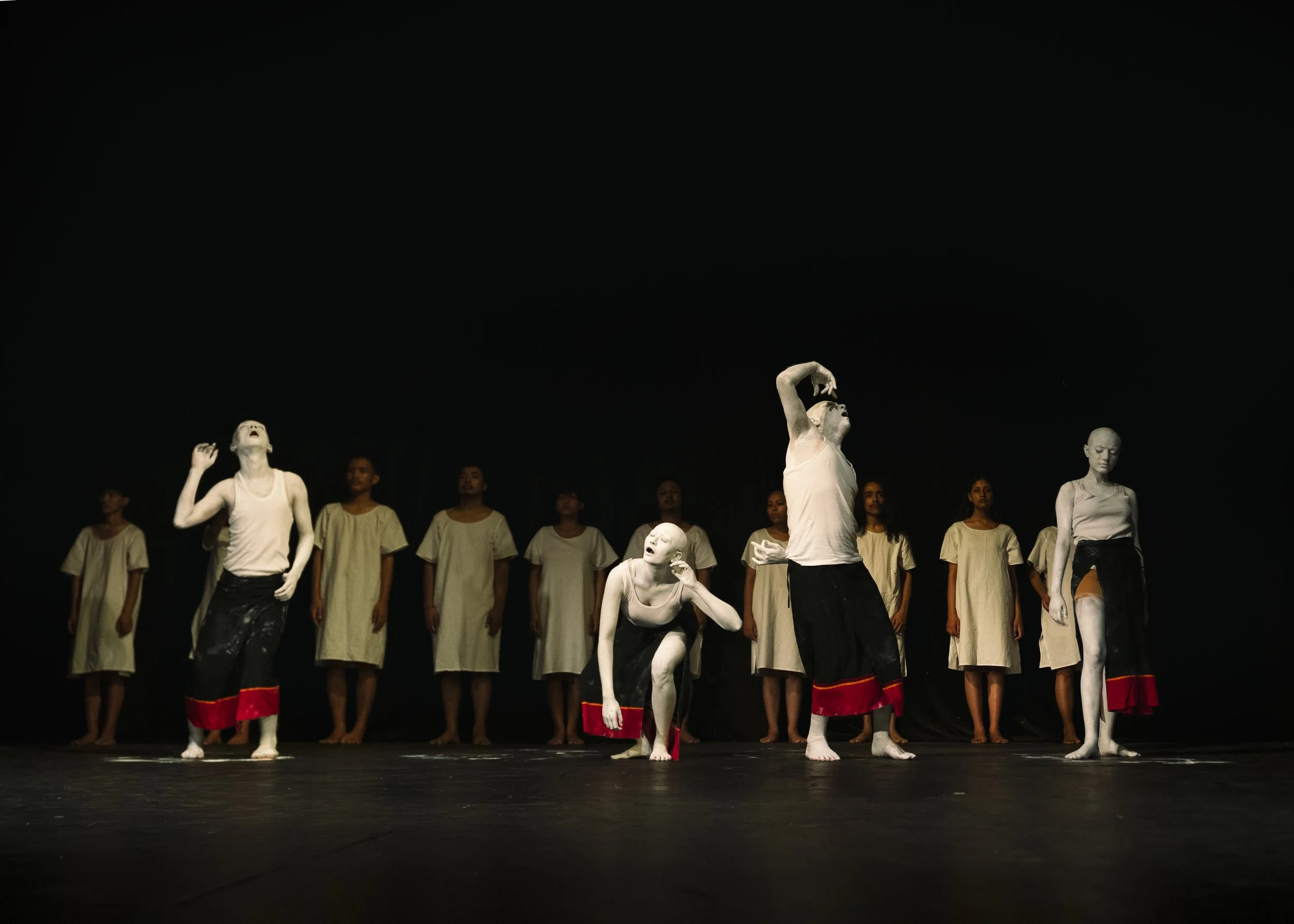 A group of performers on stage, some painted white, in theatrical costumes, with a dark background.