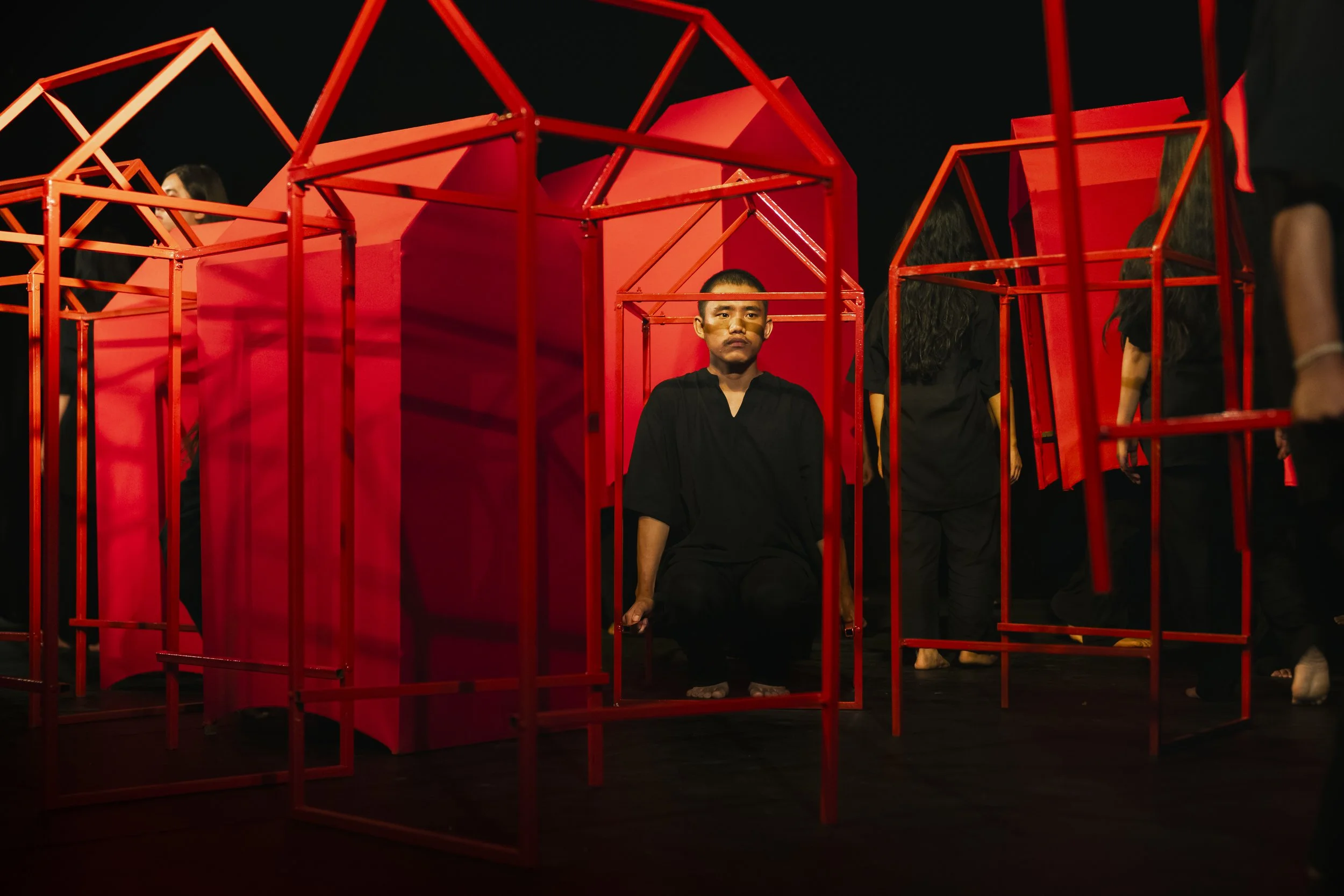 A young man in black clothing sitting inside a red, geometric cage structure on stage, surrounded by other people.