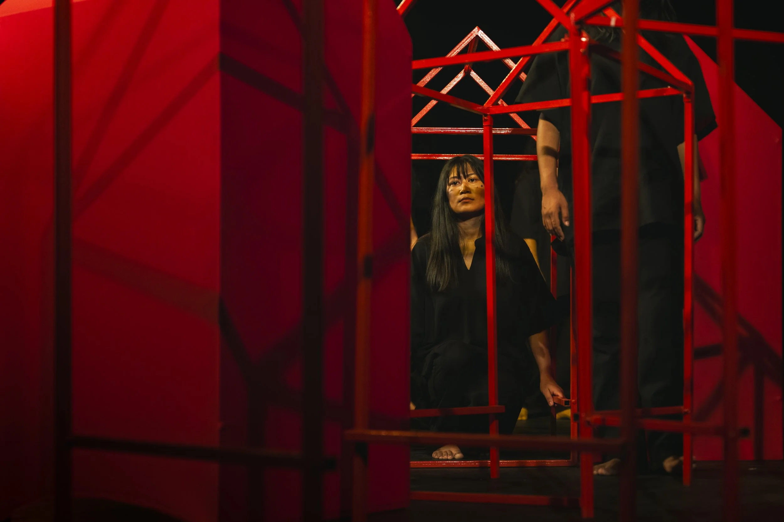 A woman sitting on the floor behind red metal frames, with black hair, wearing black clothing, and looking upward, with a dark background.