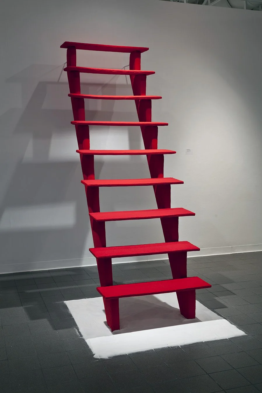 Red ladder leaning against a white wall in an art gallery with a shadow cast on the wall.