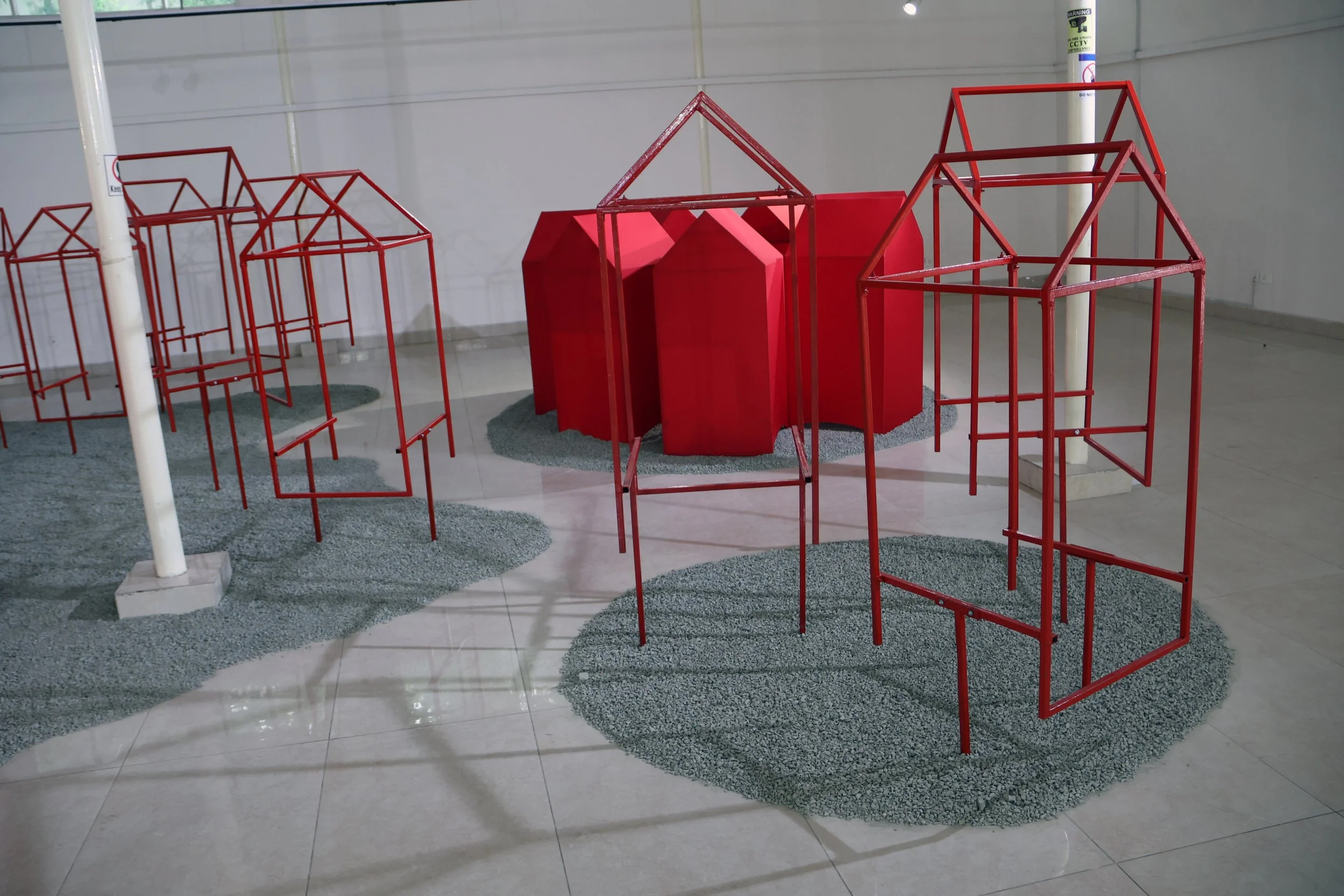Exhibition display with red metal structures resembling abstract houses or architectural models, placed on gravel-like surfaces inside an indoor space.