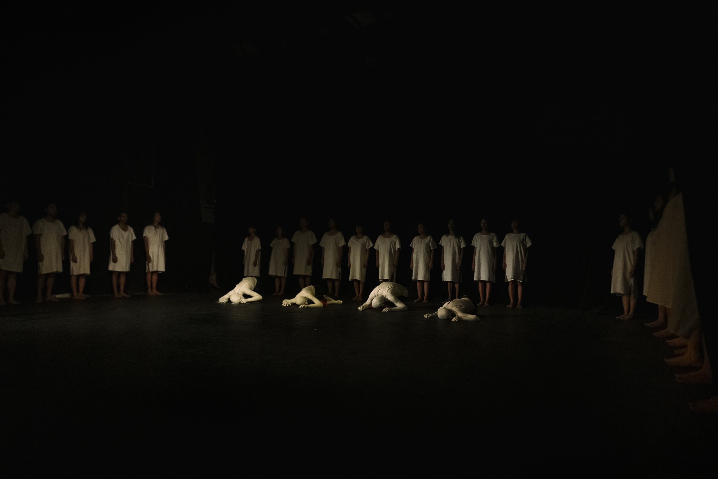 A theatrical stage scene with performers dressed in white costumes, some kneeling or lying on the floor, and others standing in a line at the back, set against a dark background.