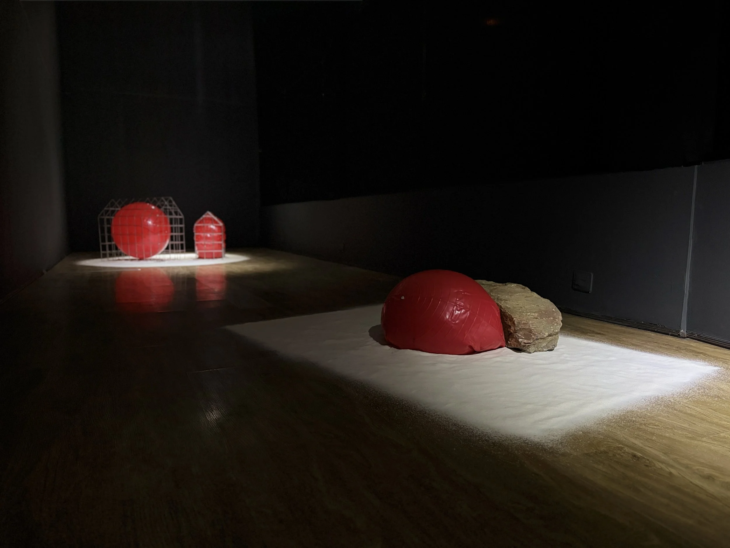 Dark room with minimal lighting, decorated with red geometric paper models, some arranged on the floor and one on a rock, illuminated by a spotlight.