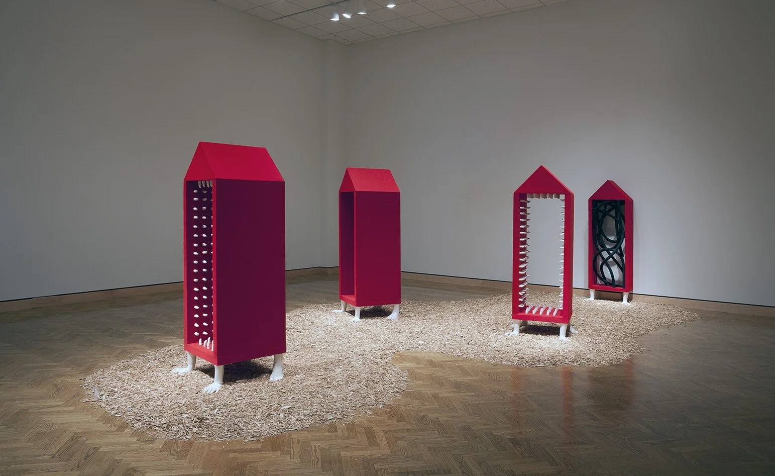 Art exhibit featuring four pink, house-shaped structures with various internal designs, displayed on wood shavings on a gallery floor.