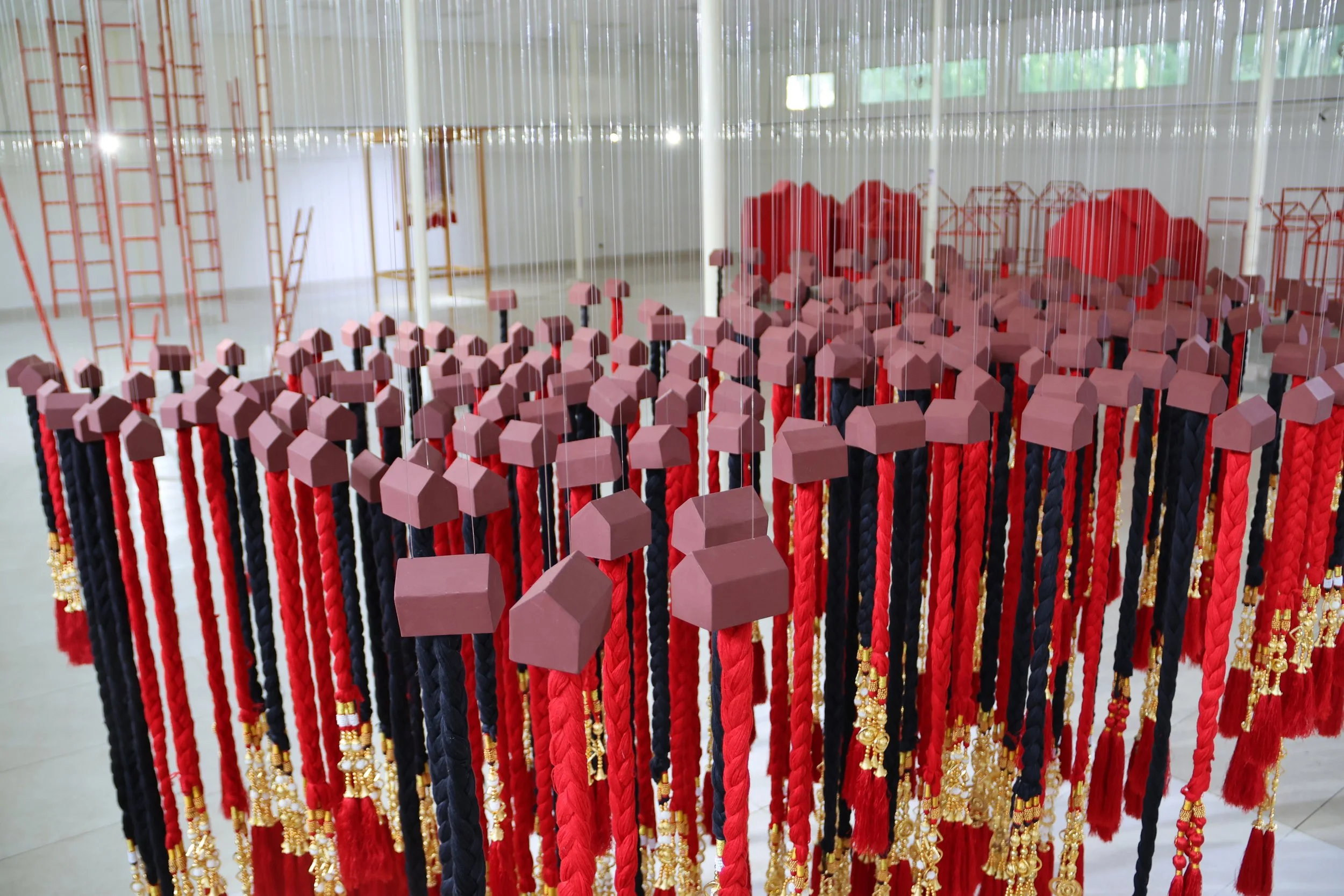 An art installation featuring numerous braided cords in red, black, and gold, with pink cubic and rectangular shapes on top, arranged in vertical rows inside a gallery space with white walls and large windows.