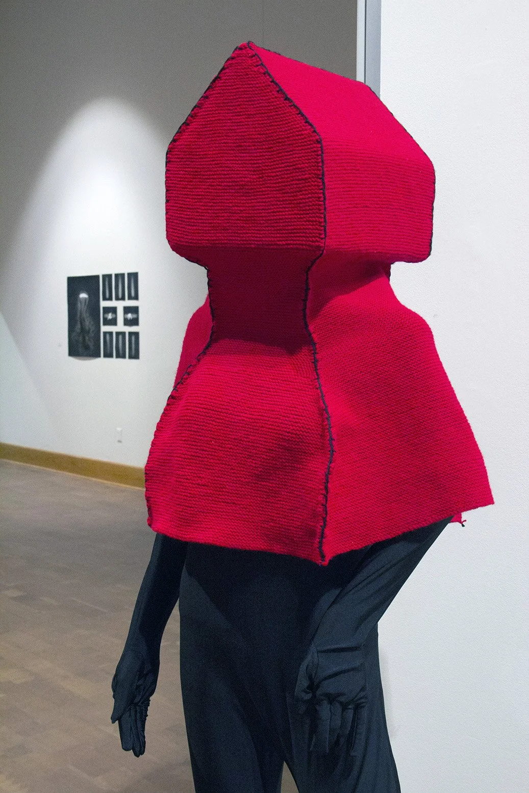 A sculpture of a humanoid figure wearing a bright pink, textured, geometric headpiece and matching top, displayed in an art gallery.