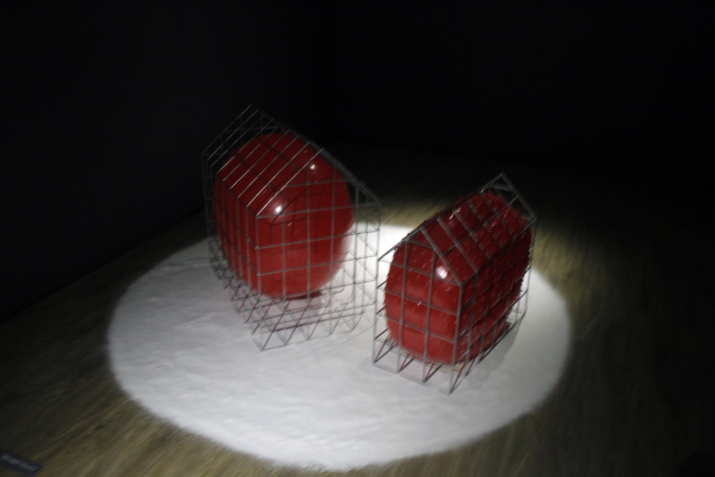 An art installation features a large red sphere inside a metal wireframe cube and an elongated red sculpture with textured bumps inside a glass display case, set on a white circular platform against a dark wall.