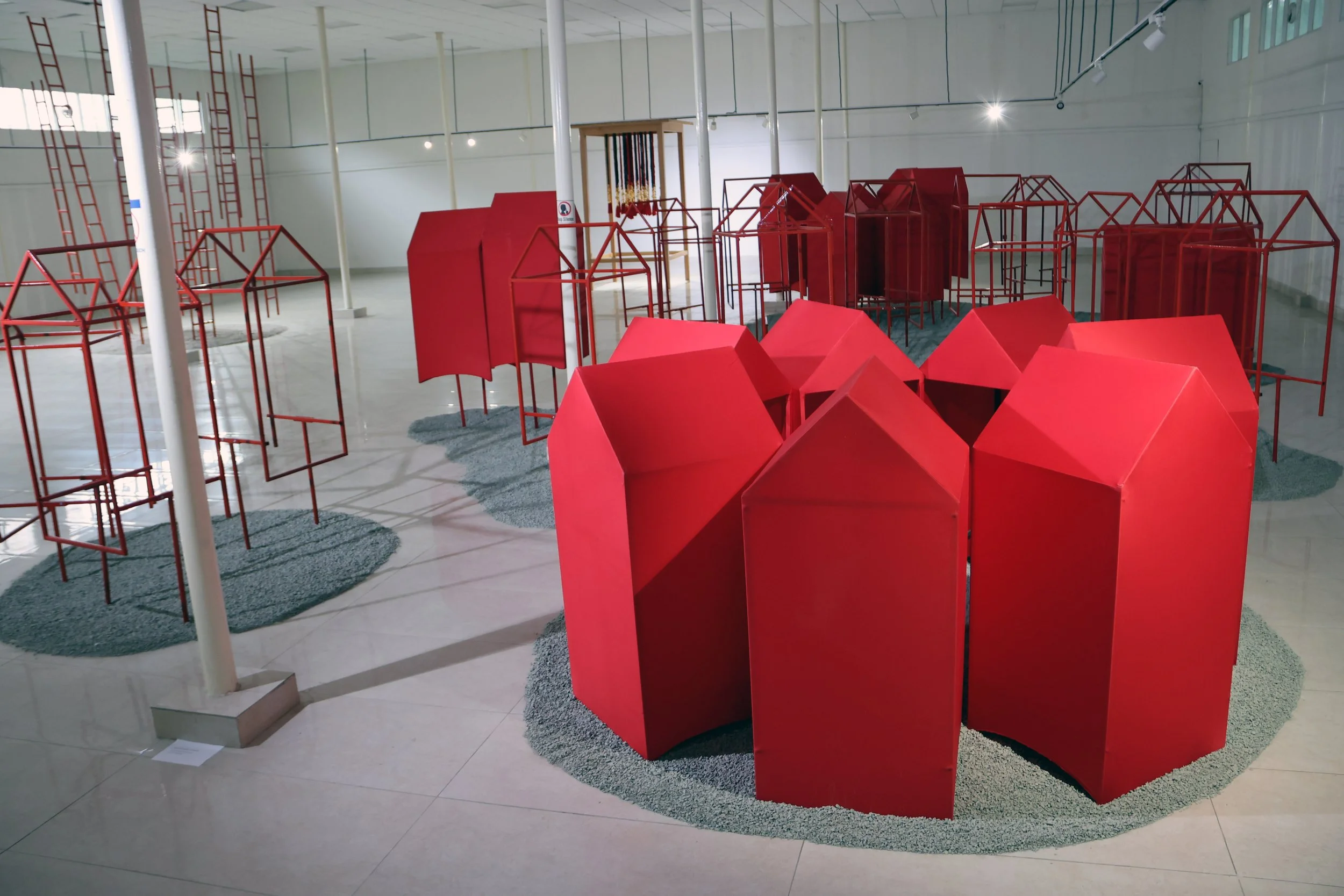 Art installation in a gallery space featuring red geometric and wireframe structures, with some structures resembling small houses and others more abstract. The space has white walls, track lighting, and a tiled floor with gravel accents around the s