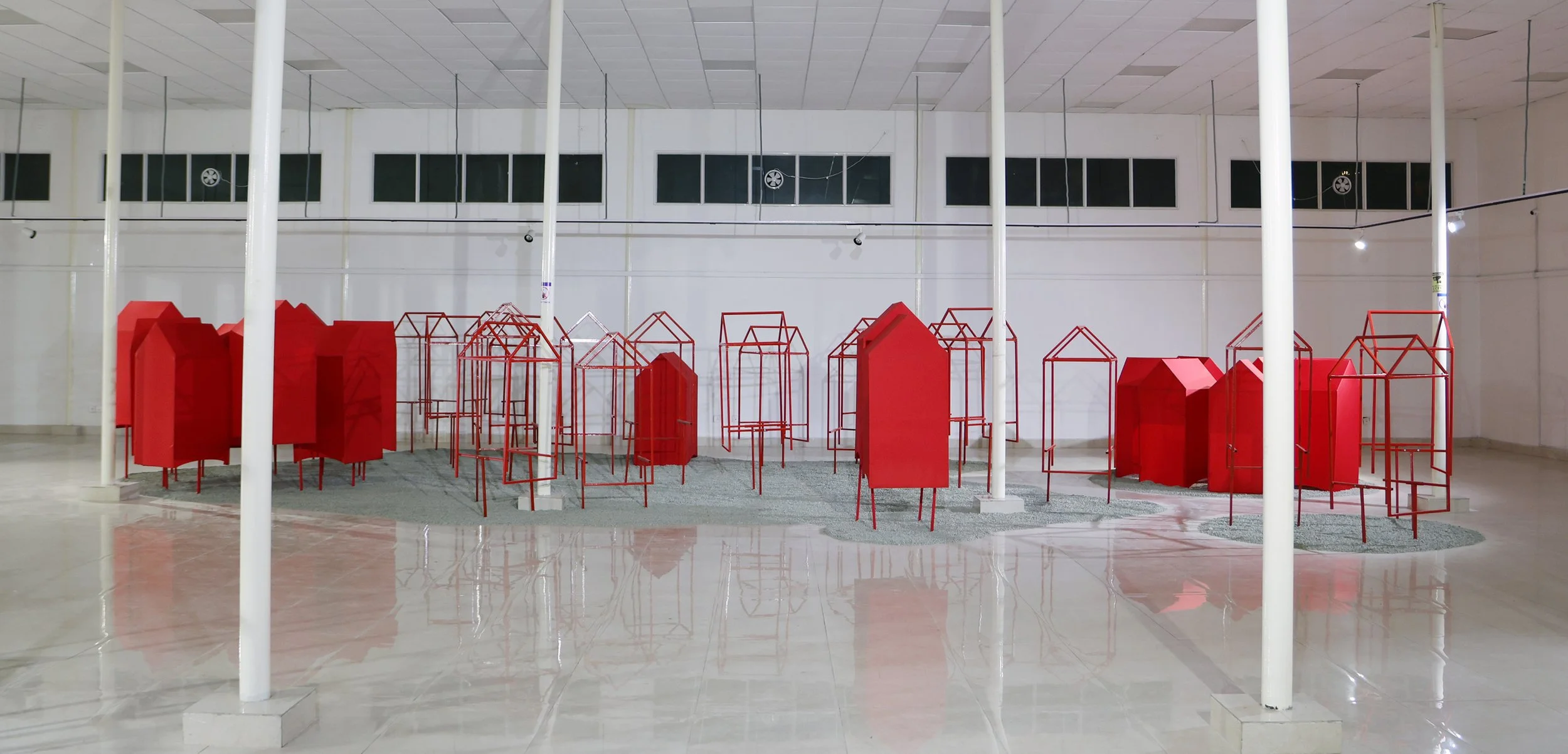 An art installation with red geometric structures resembling houses, arranged on a gray circular carpet inside a room with white walls and ceiling.