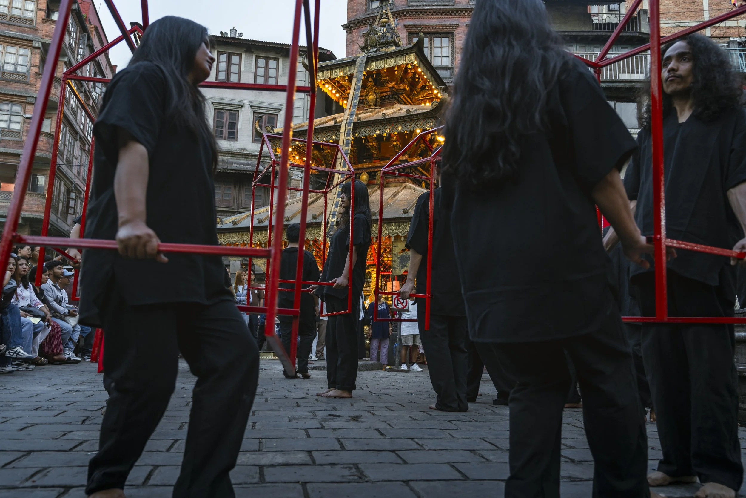 People dressed in black performing a traditional dance around a carousel in an urban setting, with seated audience and historic brick buildings in the background.