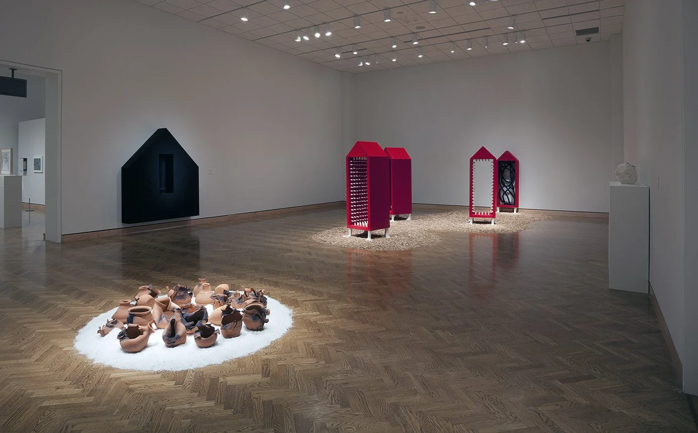Contemporary art exhibit in a gallery with various sculptures and installations, including ceramic vessels on a white rug, and house-shaped structures with different designs, displayed on a light wooden floor.