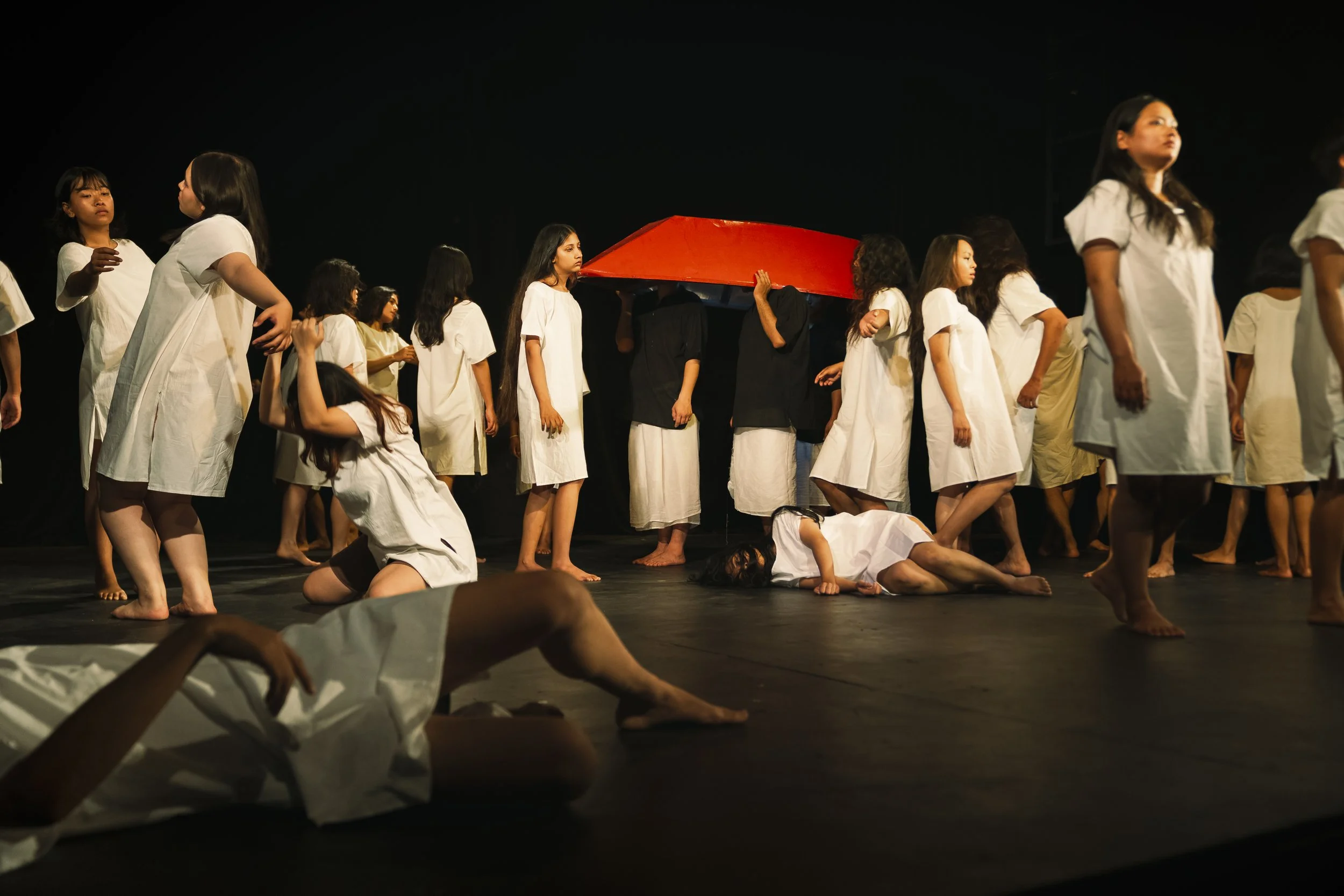Performers in white dresses perform a dance on stage, some standing, some kneeling, with neutral expressions; a person holds a large red prop in the background against a dark stage backdrop.