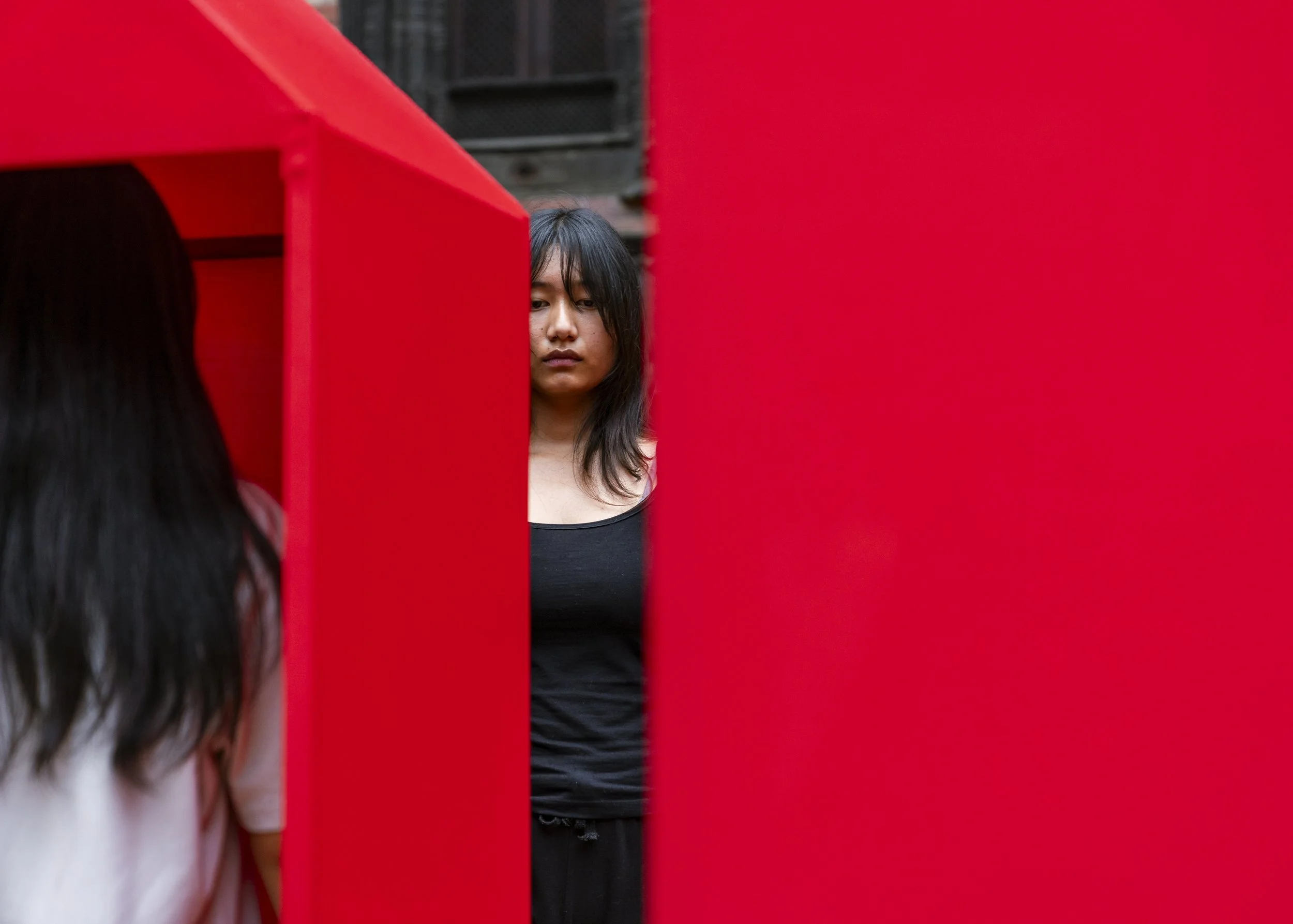 A woman with dark hair looking into a red mirror or panel, with her reflection partially visible.