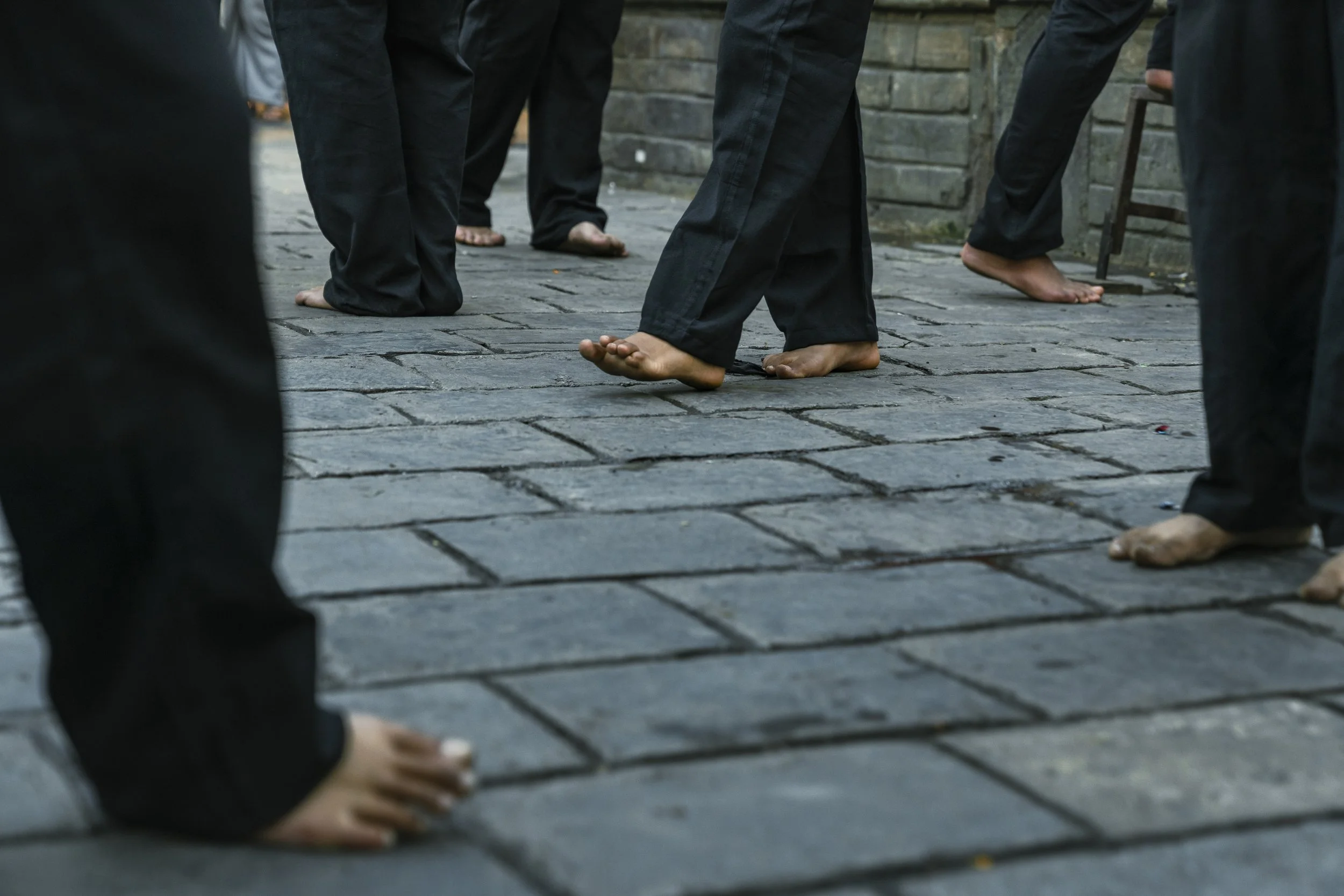 People walking barefoot on a stone pavement, with some wearing black pants and others wearing sandals or shoes.