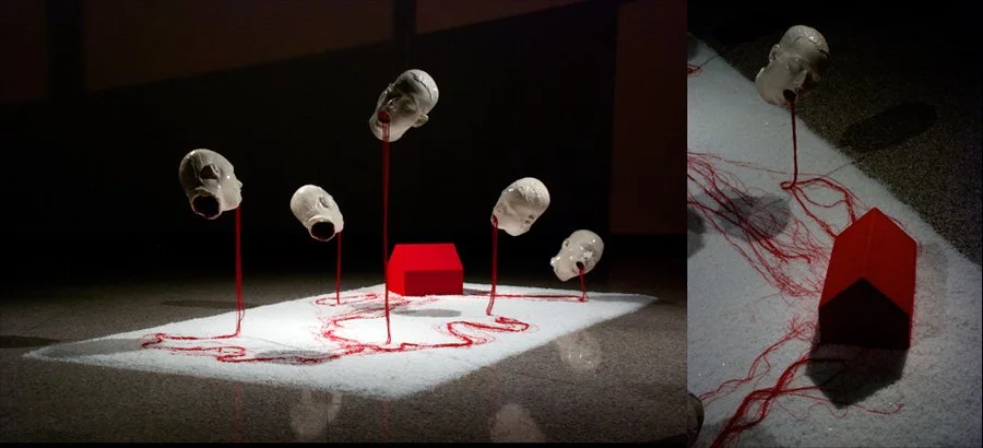 Art installation with five white mannequin heads mounted on red wires, positioned above a white surface, with red strings connecting the heads to a red box on the ground.