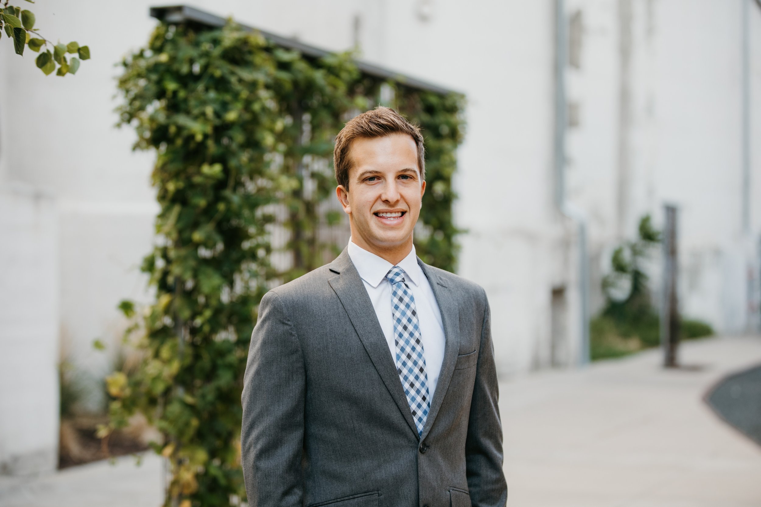 A young man in a gray suit and blue patterned tie standing outdoors near a green leafy plant, smiling at the camera.