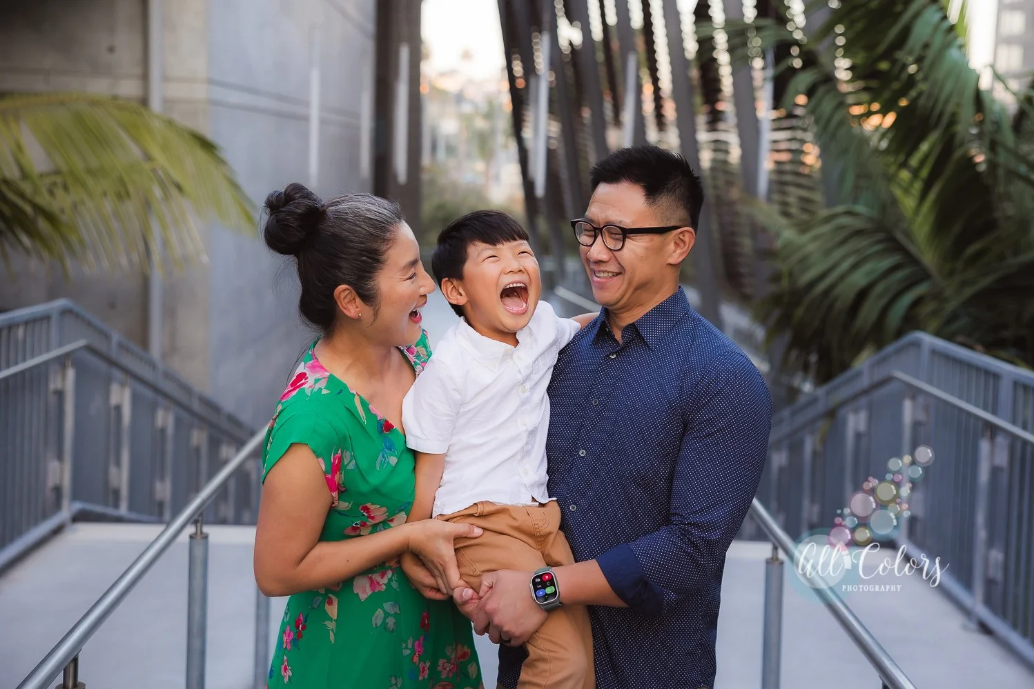 A family of three, mom, dad, and young son, smiling and laughing together outdoors during sunset. The boy is being held by the father, and the mother is holding the boy's waist, all enjoying a joyful moment on a pathway with palm trees and modern bui