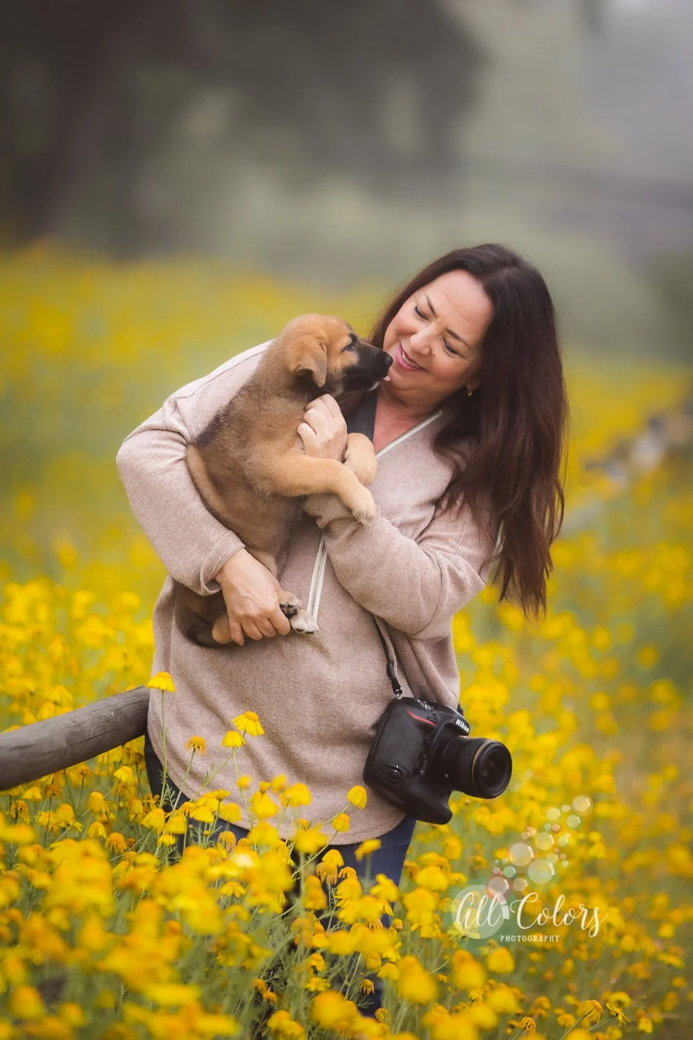 A woman holding a puppy in a field of yellow flowers, smiling and looking at the puppy.