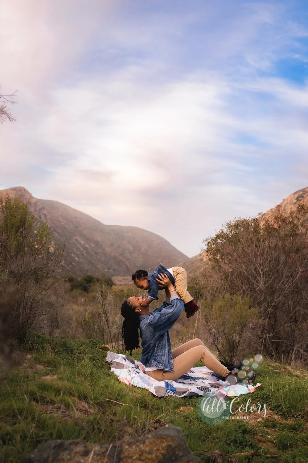 A woman sitting on a blanket in a mountainous outdoor setting, lifting a young girl into the air, with both smiling. The sky is partly cloudy with a mix of blue and pink hues, and there are bushes and mountains in the background.
