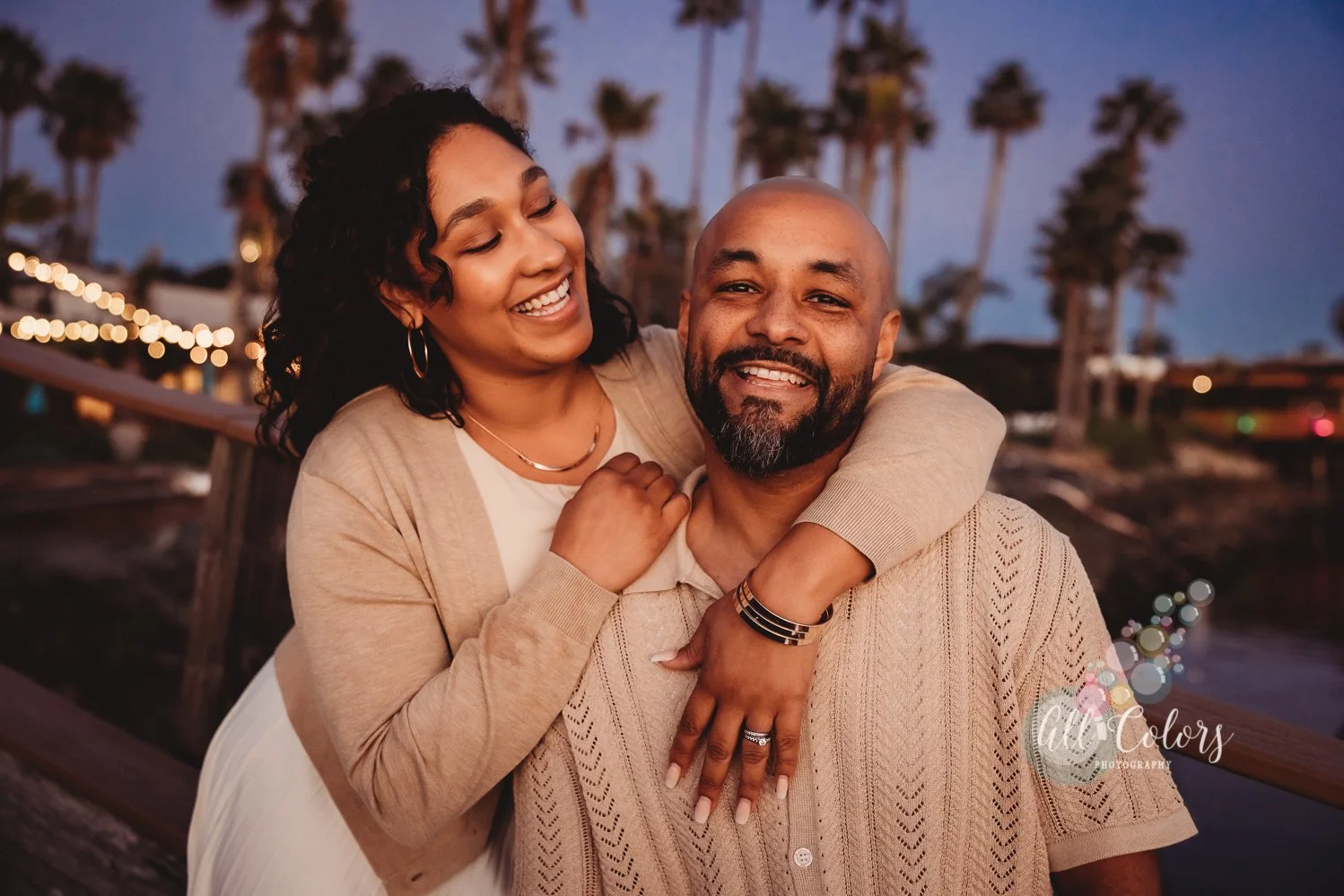 A happy couple enjoys a moment together outdoors during sunset, with palm trees and string lights in the background.