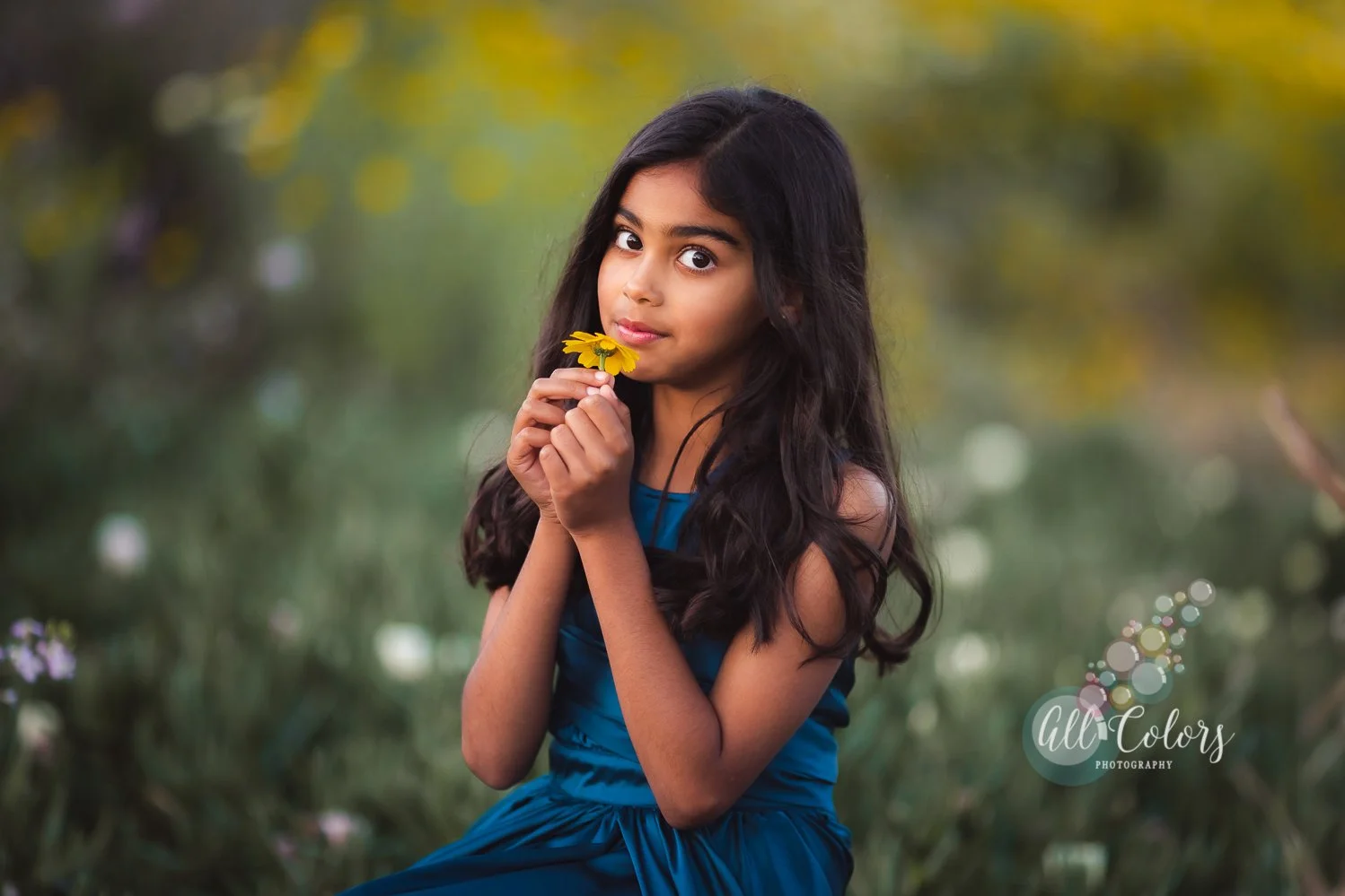 Young girl with long dark hair wearing a blue dress holding a yellow flower to her face outdoors.