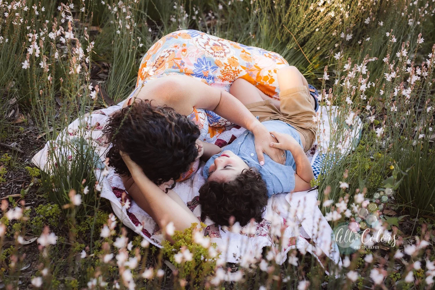 A woman and a young boy lying on a blanket in a field of white flowers, cuddling and looking at each other.