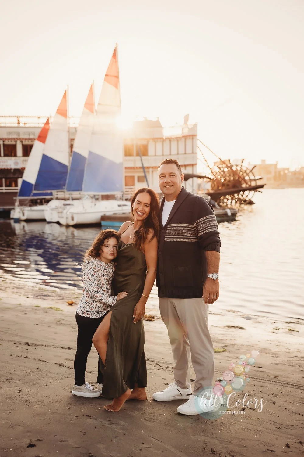 A family of three standing together on a sandy beach at sunset, with sailboats and a dock in the background.