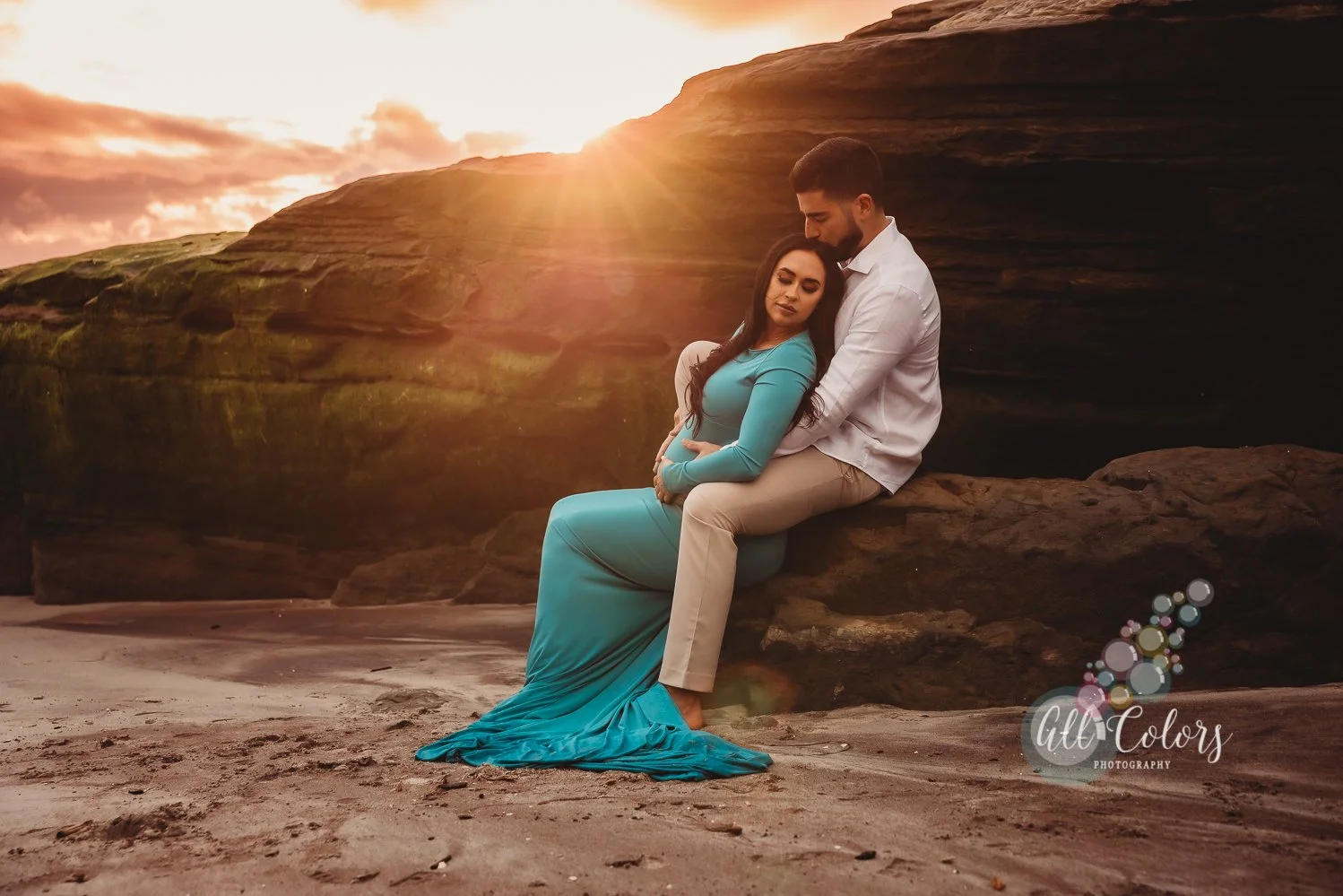 A couple sitting on a rock at the beach during sunset, with the man embracing the woman as she leans into him. The woman is wearing a long turquoise dress, and the man is dressed in a white shirt and beige pants.