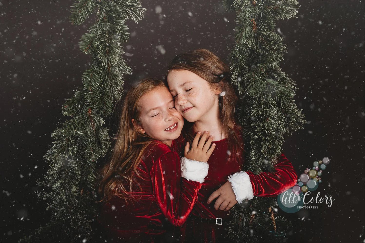 Photo of two sisters dressed in red velvet Christmas outfits on a winter swing with snow falling around them.
