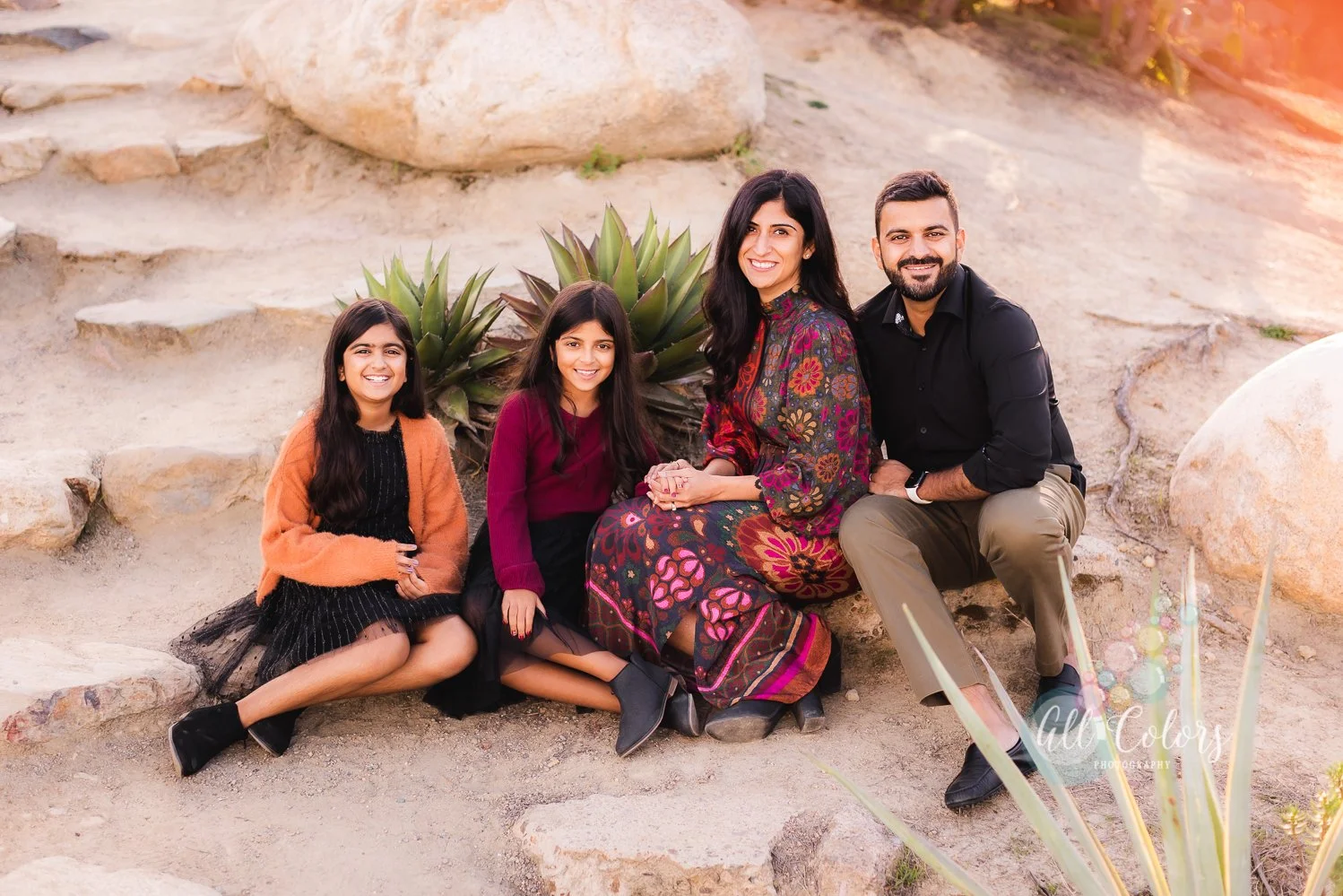 Family of four sitting on the ground of a desert garden in san Diego looking and smiling at the camera.