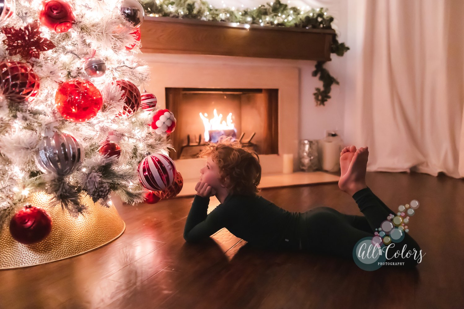Boy with curly blonde hair laying on the wood floor looking at a lit up Christmas tree