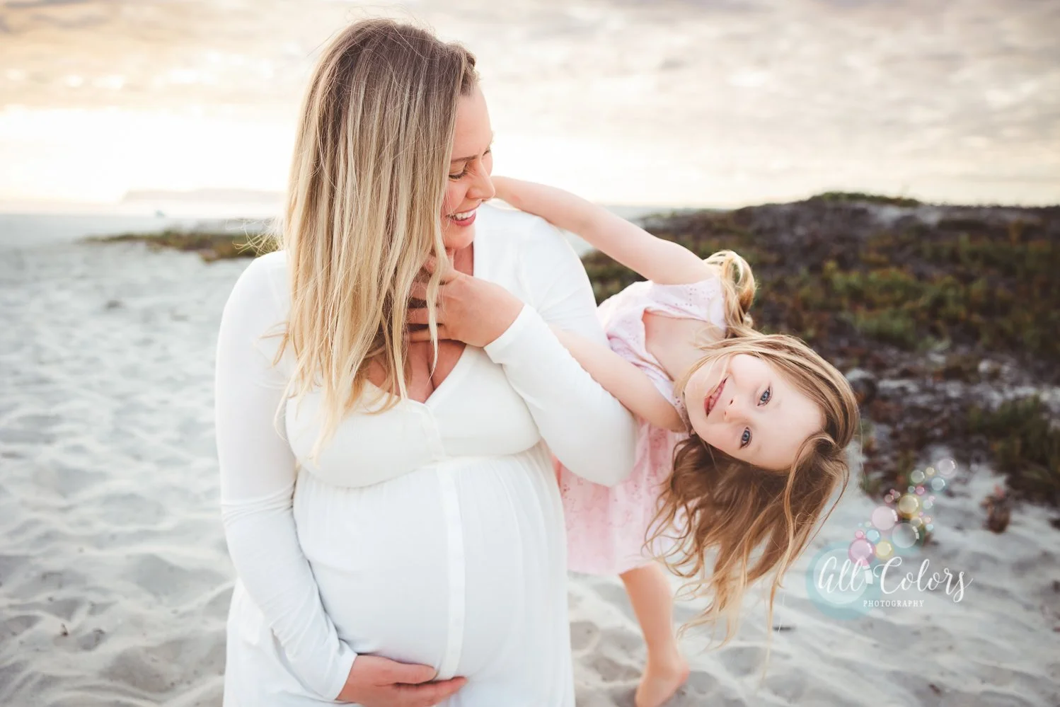 Expecting woman wearing a white dress plays with young daughter at the beach