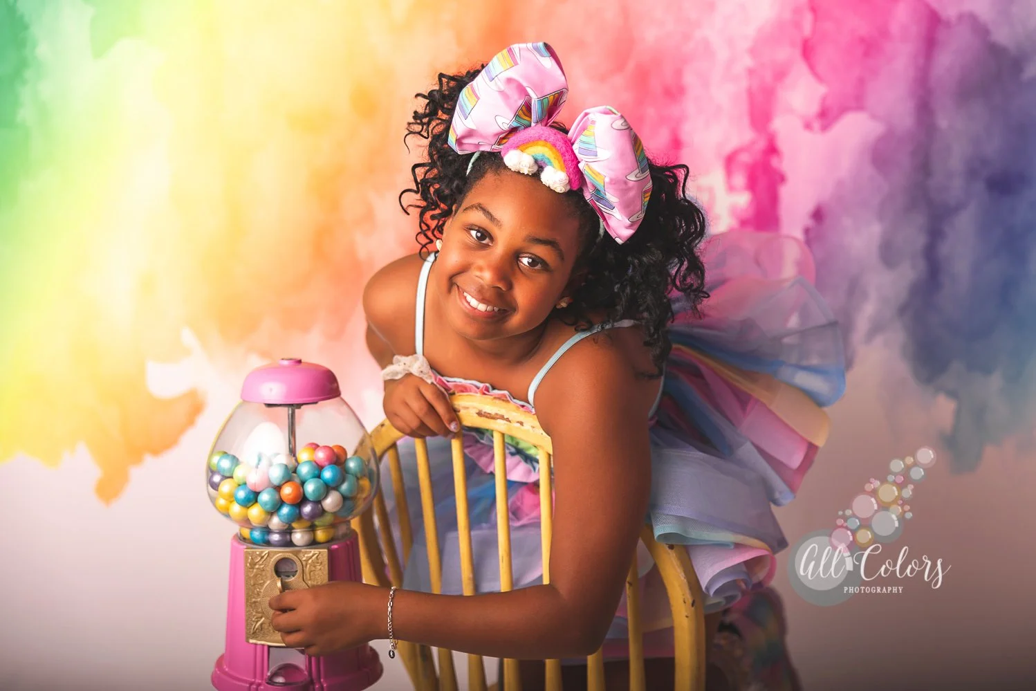 Black girl wearing a huge rainbow bow in front of a very colorful backdrop for a birthday session