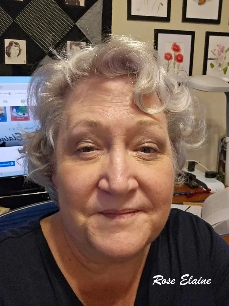 A woman with short, curly gray hair taking a selfie in an office or art studio, with framed artwork and a computer screen in the background, and the text 'Rose Elaine' in the bottom right corner.